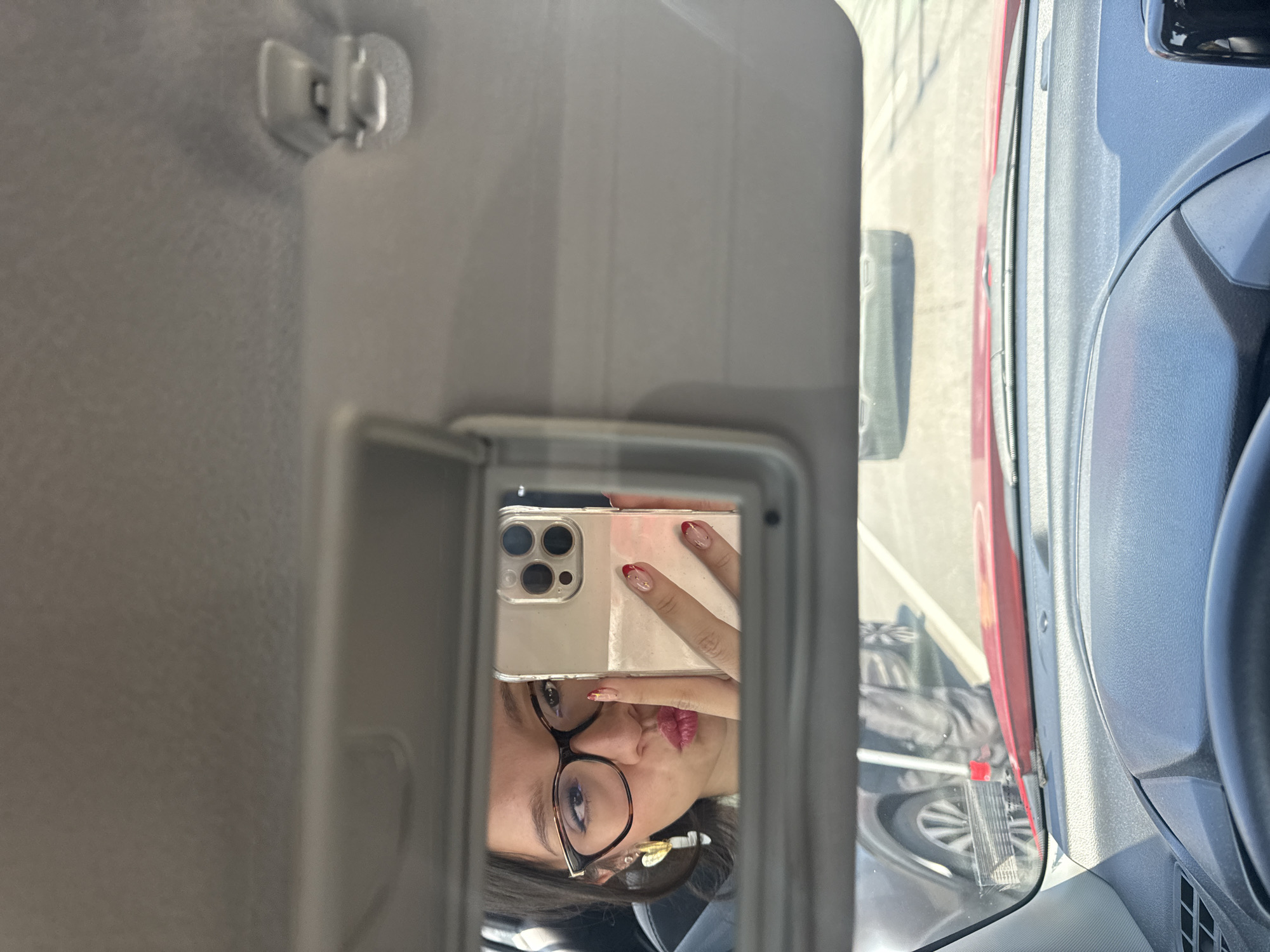 The image, oriented vertically, depicts a person taking a mirror selfie from inside a car in San Andrés Cholula, Mexico. The central focus is the person's reflection in the car's passenger-side visor mirror. The individual, wearing dark-rimmed glasses, has dark hair and red lipstick. Their hand, holding a smartphone with a clear case, is visible, showcasing nails with a red-tipped French manicure. The smartphone, likely an iPhone, features a prominent triple-lens camera system. A golden hair clip or earring can be seen near the person's ear.
The car's interior, including the textured grey sun visor and headliner, frames the mirror on the left. To the right of the visor, through the car window, an outdoor scene is visible. A bright red vehicle, possibly a truck or large car, is parked closely alongside. Faint details of another vehicle's wheel and tire are also discernible in the background through the window or reflection. The bright, natural lighting indicates it is daytime. No discernible text is visible in the image.