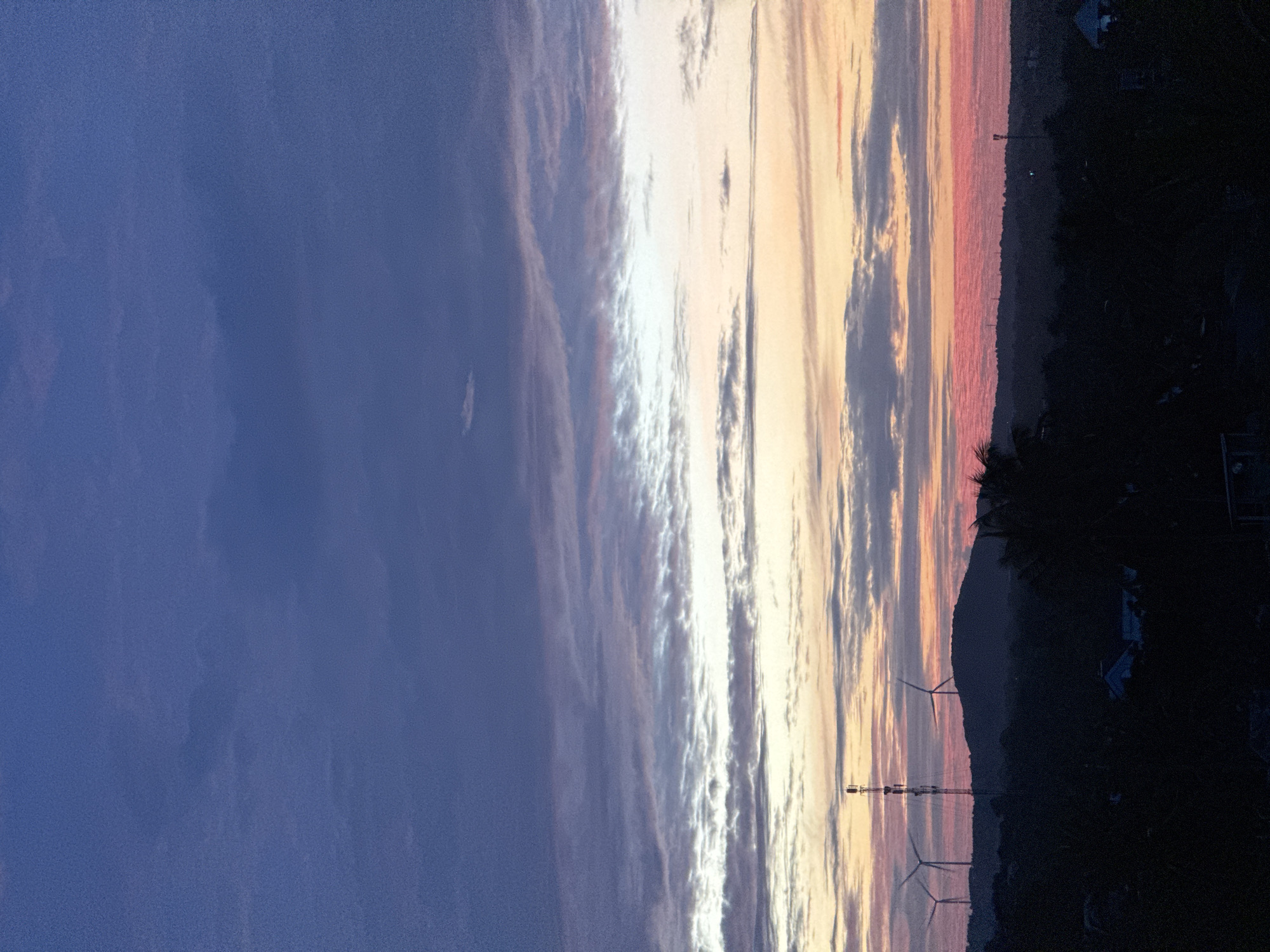 A low-angle, vertical shot captures a dramatic sunset over Mũi Né, Vietnam. The sky transitions from a deep indigo at the top to bands of purple, pink, and orange closer to the horizon, painted with wispy clouds. Silhouetted against this vibrant sky are rolling hills dotted with wind turbines and a tall telecommunications tower. The foreground is a dark, dense landscape of trees and rooftops, suggesting a residential or rural area within the city. There are no people visible in the image. The overall impression is one of natural beauty and the presence of renewable energy infrastructure.