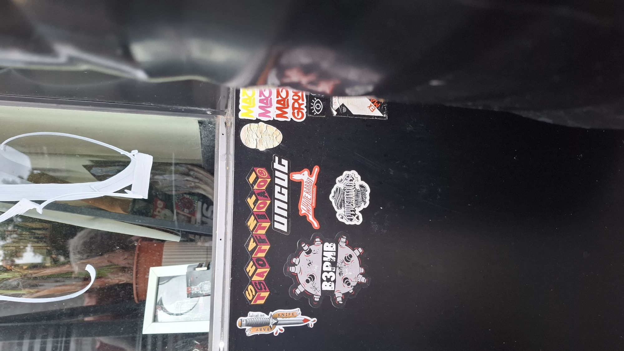 The image displays a close-up view of stickers on a dark, possibly metal surface, which could be a vehicle or a piece of street furniture. The stickers are varied in design and content. One prominent sticker shows a red and white illustration of a stylized knife with blood dripping from the blade. Above this, a series of yellow cube-shaped stickers with red lettering spell out "TSAOTFIOAO". Below the knife sticker, a circular sticker features a grey, spiky object resembling a mine, with the Cyrillic word "ВЗРИВ" (which translates to "explosion") in large white letters across it. To the right of these, a red sticker depicts a white rabbit in mid-leap with the words "WHITE RABBIT" underneath. Further to the right, a cluster of vertical stickers show the word "MAZ" repeated in different colors (yellow, pink, red) followed by "GROO" and an eye symbol. There is also a sticker with a textured, light-colored face and a white sticker with stylized lettering that appears to say "UNCUT". Another sticker, to the lower right, is a white, somewhat distressed design with black lettering.
In the background, visible through a window or reflective surface, are reflections of indoor elements. A potted plant with long, thin leaves is visible, along with what appears to be a white framed picture or mirror. The overall setting suggests an urban environment, possibly a street or a public area in Plovdiv, Bulgaria, given the Cyrillic script on one of the stickers. The surface with the stickers is the primary focus, suggesting it is the subject of interest. There are no people clearly visible in the image.