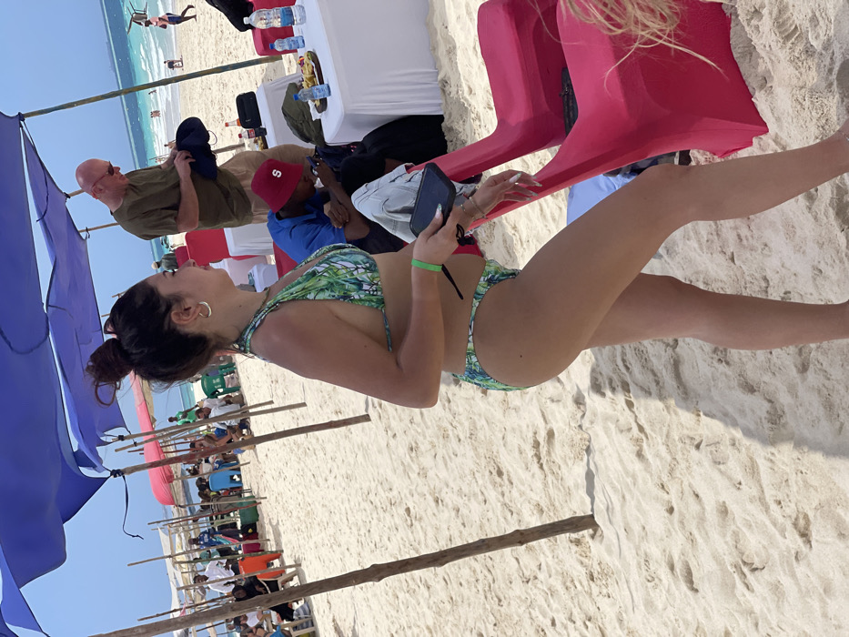 A sunny beach scene on a white sand beach in Zanzibar, Tanzania, under a clear blue sky.
In the foreground, a young adult female with dark hair tied in a bun stands in profile, facing right. She wears a two-piece green and white tropical print bikini and a neon green wristband on her left wrist. She holds a black smartphone in her left hand, displaying white fingernails. Her right arm is partially obscured.
In the midground, under blue canopies supported by wooden poles, several individuals are present. To the right, a bald adult male in an olive green shirt and beige shorts stands behind a white-tablecloth-covered table, looking down at and handling a dark object, possibly a hat. Seated at the same table is a dark-skinned individual wearing a blue shirt and a red bucket hat with a white 'S' logo, looking down at a smartphone. The table holds several plastic water bottles, two soda bottles, and a wooden tray with fried snacks. Two bright pink fabric-covered chairs are positioned near the table. Further left, other individuals are visible under blue and red canopies, some seated at tables, others standing or walking. Figures include men in white shirts and a person in an orange garment.
The background reveals a turquoise ocean with whitecaps and additional people wading or swimming. More individuals are scattered along the beach near the waterline. The overall environment is a bright, clear day at a tropical beach.