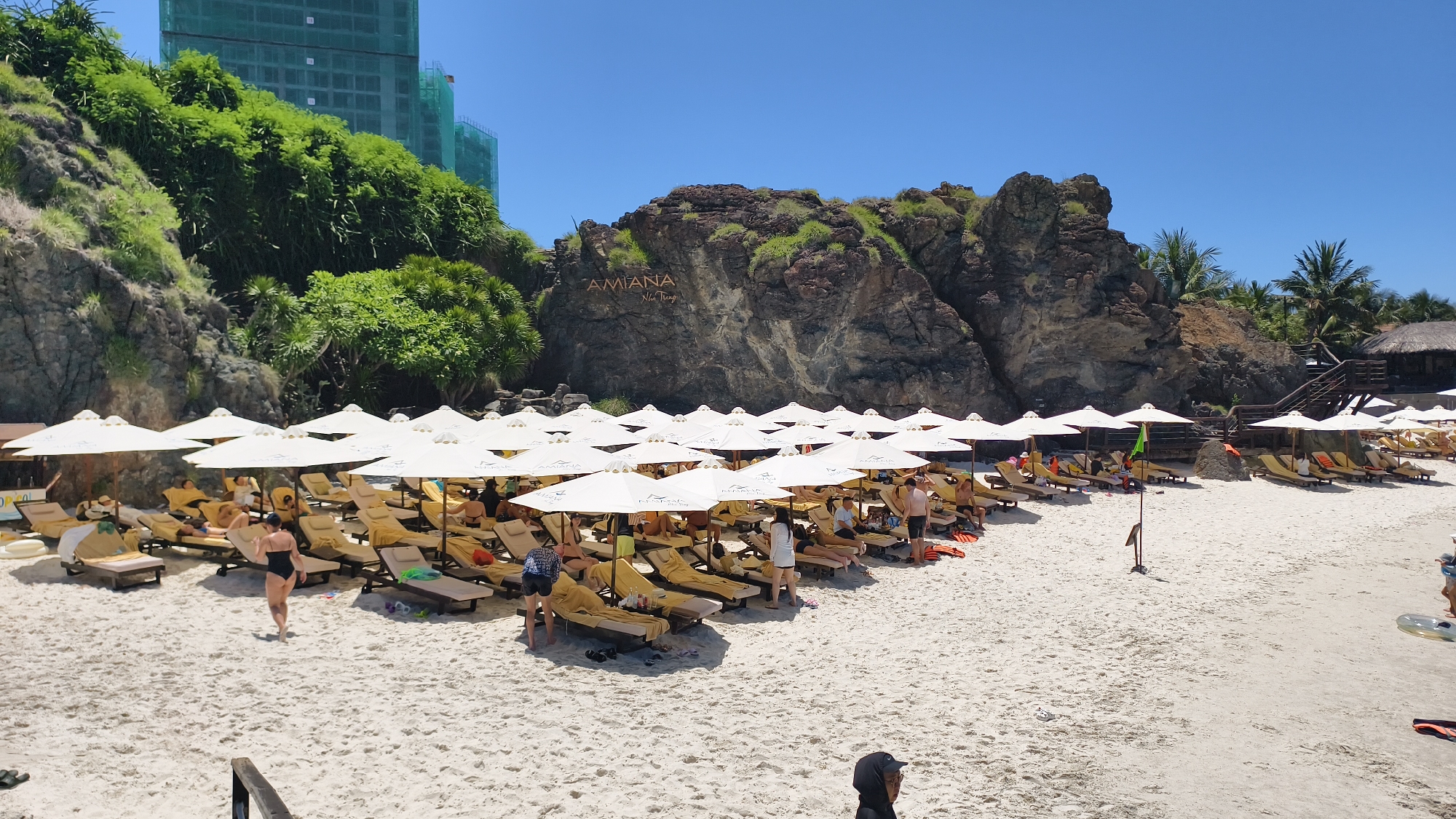 A wide shot captures a crowded beach on a sunny day, with many people relaxing on lounge chairs under white umbrellas. The beach is covered in white sand, and lush green vegetation covers the rocky cliffs in the background. Two large, unfinished buildings, covered in green netting, are visible behind the cliffs. The "AMIANA Nha Trang" logo is visible on the cliff face. Several individuals are depicted in swimsuits, some are lying on lounge chairs, while others are standing or walking on the sand. In the foreground, a dark-hooded person is partially visible. The sky is clear and blue.