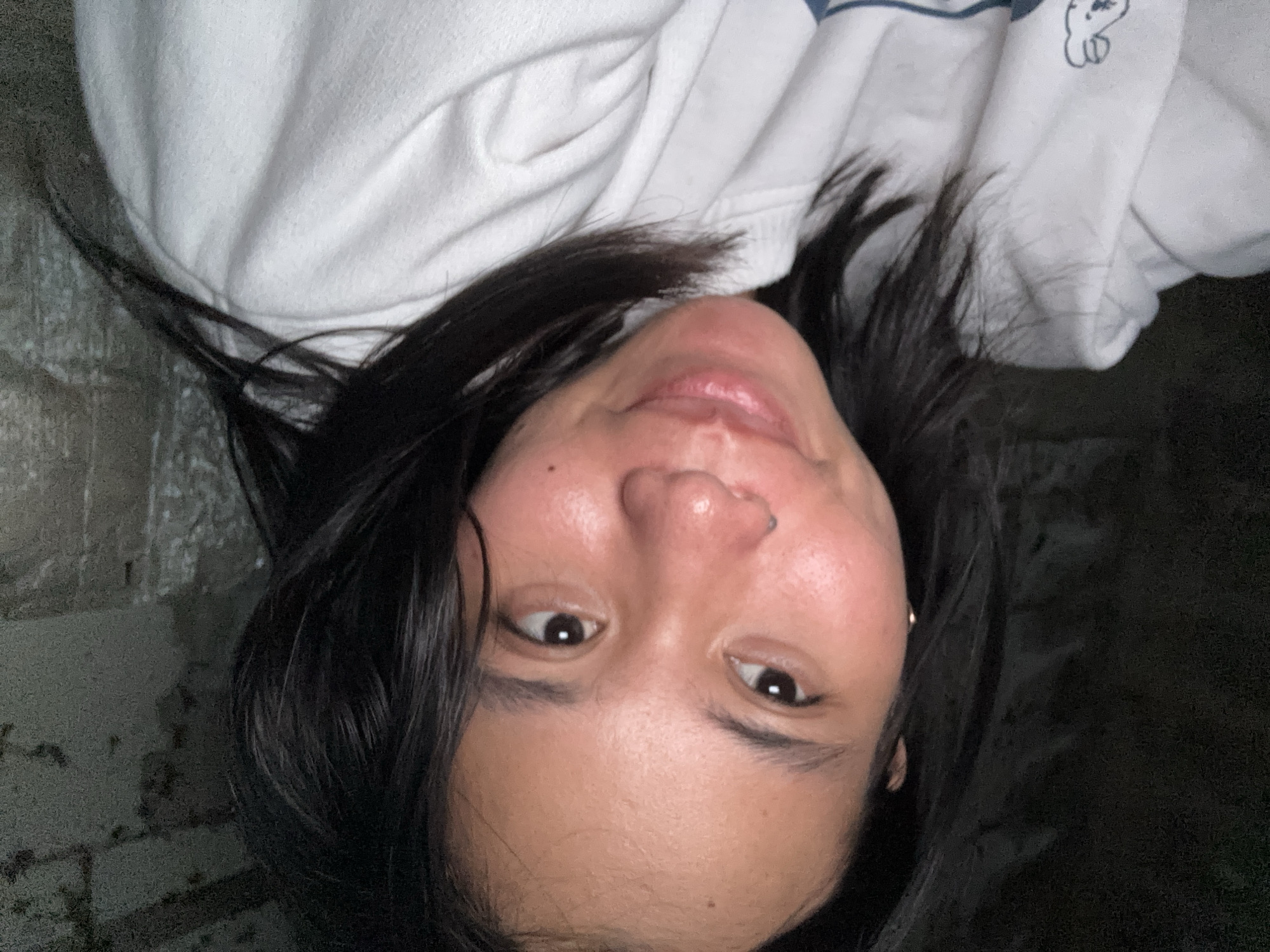The image shows a close-up, selfie-style shot of a young East Asian woman with dark hair and brown eyes. She is wearing a white sweatshirt. The background appears to be a rough, concrete wall. The lighting is dim, creating a moody atmosphere. No specific location in East Jakarta is identifiable from the image.