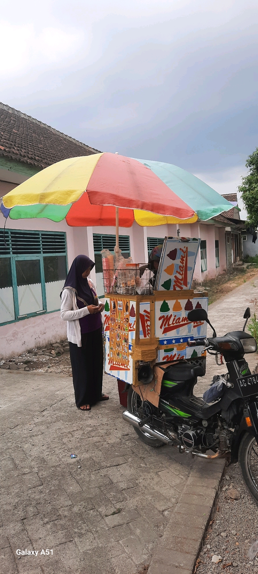 In Tangkil, Indonesia, an outdoor scene captures an ice cream cart on a paved pathway. A woman wearing a dark hijab and a light-colored cardigan over a purple top stands beside the cart, looking at her phone. The ice cream cart is mounted on a black motorcycle with green accents. The cart itself is decorated with images of ice cream cones and the word "Miami." A colorful umbrella, with segments of yellow, pink, and light blue, is propped open above the cart. In the background, a light pink building with green-framed windows is visible, situated behind a tiled roof. The sky overhead is overcast.