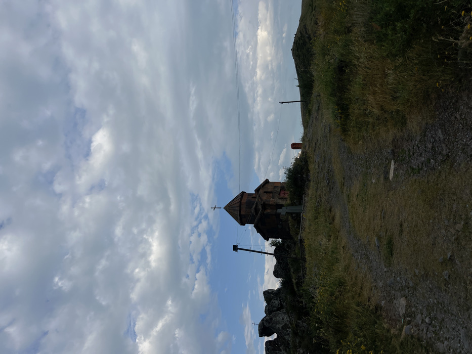 A stone church with a cross sits on a hillside, against a backdrop of cloudy skies. A dirt path winds alongside the church, lined with grass and rocks. Power lines stretch across the sky. The angle makes the scene appear skewed, with the sky on the left and hillside on the right. The architectural style of the church is traditional.