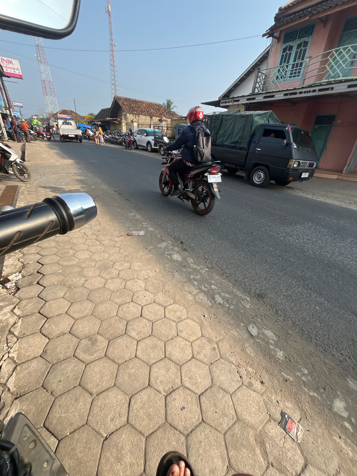 The image depicts a street scene in Kemuning, Indonesia, under a clear blue sky. In the foreground, a motorcycle handlebar is visible from the rider's perspective, with hexagonal paving stones on the left side of the road. A black pickup truck with a green tarp covering its cargo bed is driving on the asphalt road. To the right of the truck, a pink building with light blue window frames and a balcony can be seen. A person wearing a red helmet and a backpack is riding a red and black motorcycle in front of the pickup truck. Other motorcycles, a white pickup truck, and people are visible further down the street, along with some shops and communication towers in the background. The overall impression is of a typical day in a busy street in an Indonesian town.
