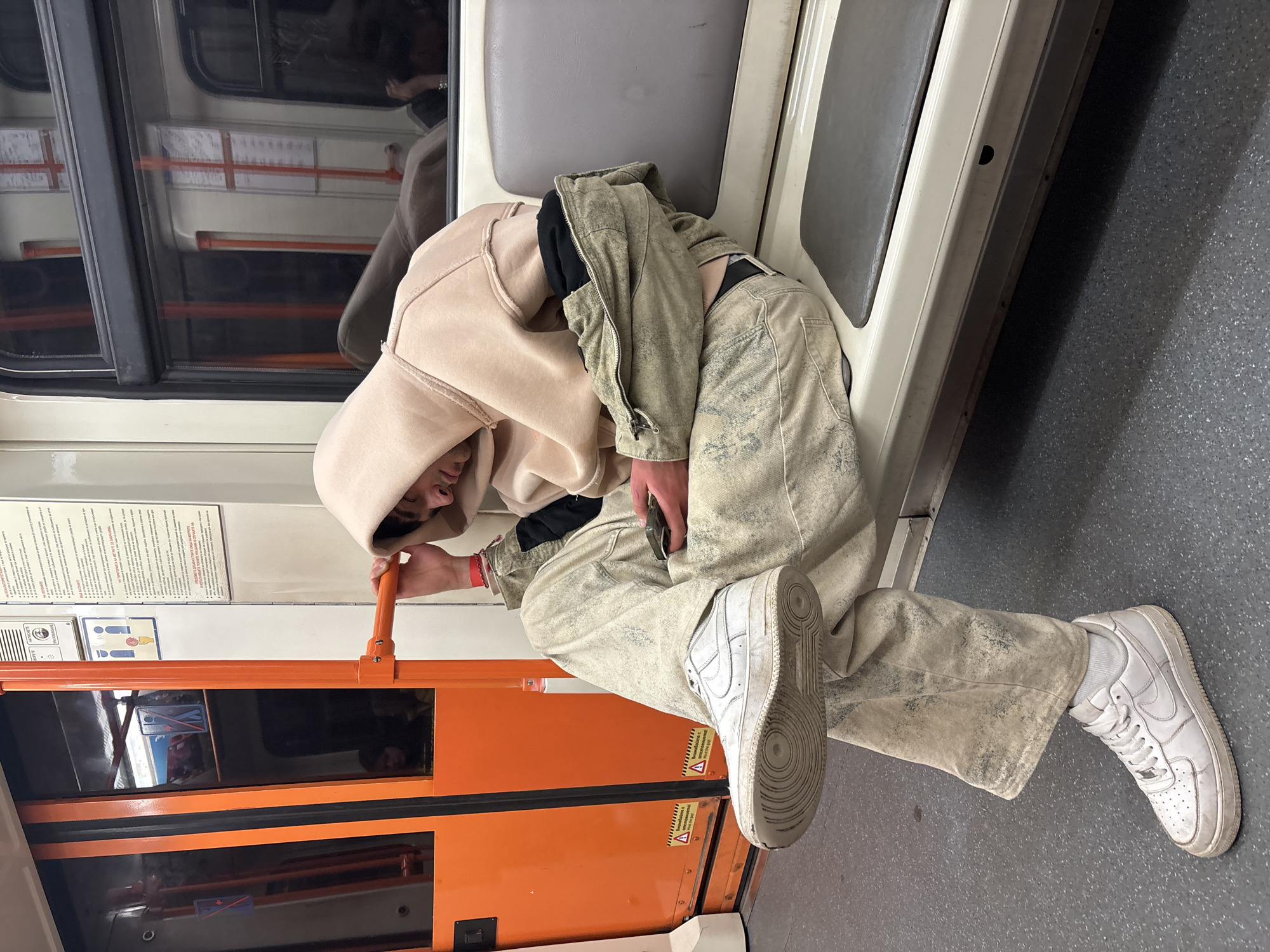 A person is sitting on a seat inside what appears to be a subway car, possibly in Sofia, Bulgaria, judging by the orange door of the train car. The person is wearing a beige hoodie with the hood up, tan pants, and white sneakers. The person's head is resting near the window, and they are holding onto a vertical orange pole. The background shows a window and interior of the subway car.