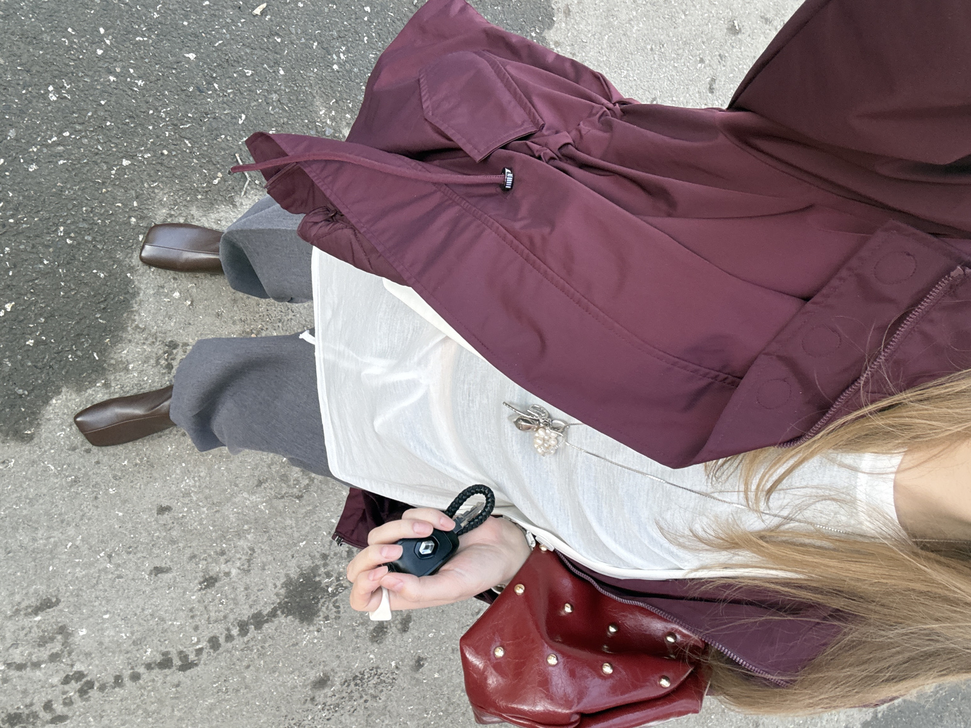 A high-angle shot shows a person standing on a concrete surface, likely a sidewalk, based on the visible texture. The person is wearing grey pants and brown shoes. Over the white top is a burgundy-colored coat or jacket. The person is holding a car key in their hand. A red purse is visible near the hand holding the key. A necklace is draped over the white shirt. The location is assumed to be Izhevsk, Russia, but without specific landmarks or contextual clues in the image, it cannot be verified.