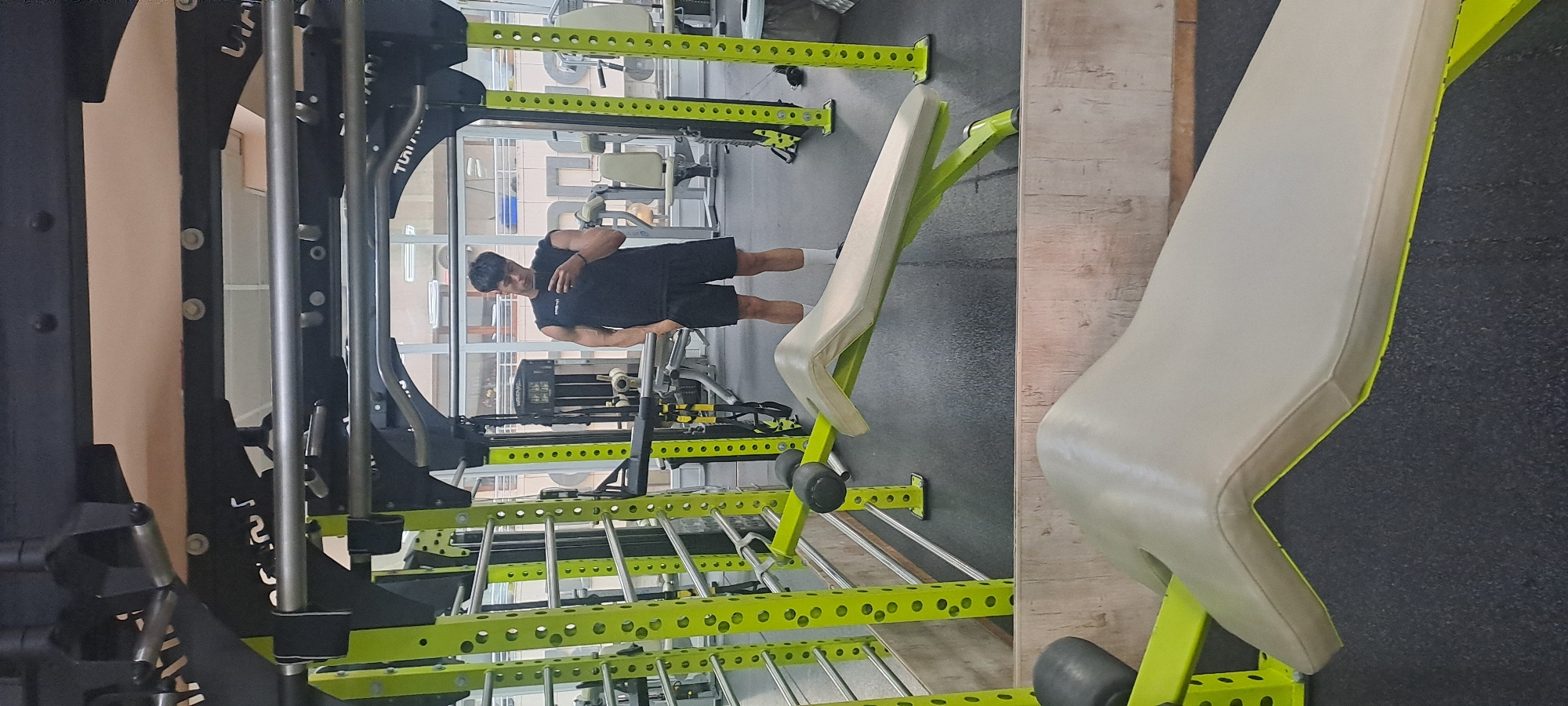 The image, originally rotated 90 degrees clockwise, captures an indoor gym scene in Burgas, Bulgaria.
The primary subject is a man, appearing to be in his 20s or 30s with short dark hair, taking a selfie. He is dressed in a black sleeveless top, black shorts, and white socks, holding a smartphone in his right hand to capture his reflection. He stands upright, facing the camera, positioned in front of various gym equipment.
The setting is a modern fitness center, characterized by large, robust gym equipment. A prominent feature is a multi-functional gym rack with a strong black frame and distinctive bright lime green crossbars and safety arms, which feature numerous adjustment holes. Chrome-plated bars are also part of this structure. Adjacent to the rack, a light-colored (beige or off-white) gym bench with a matching lime green frame is visible. A small dumbbell rests on the floor near the rack. The background reveals reflections of other gym equipment and elements, suggesting the presence of mirrors or large windows.
Visible text includes partial branding on the black frame of the gym rack, which appears to be "LINIT" and a reflected, inverted version that might read as "TUIITIL" or similar, indicating a brand name. The overall lighting suggests it is daytime. The man's expression is neutral and focused on taking the picture.