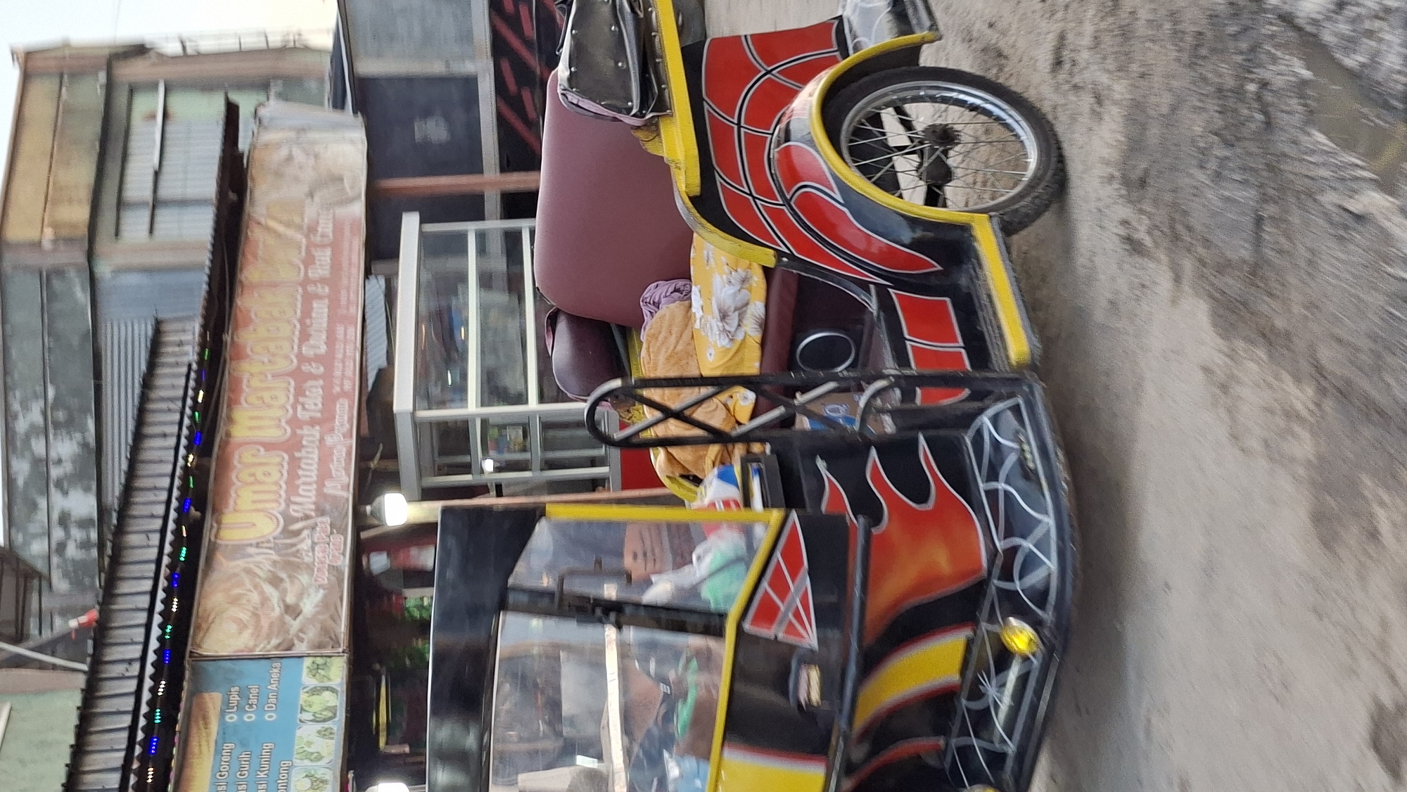 A side view of a brightly colored, three-wheeled vehicle, possibly a modified motorcycle or auto-rickshaw, is parked on a dirt road in Keudegeudong, Indonesia. The vehicle is predominantly yellow and black, with red flame-like designs on its body. It has a cushioned seating area, appearing to be maroon and burgundy, covered with blankets and what looks like a rolled-up cloth. The vehicle has a metal frame for protection, and a single wheel is visible on the right side. In the background, a building with a corrugated metal roof is visible, with a banner displaying "Umar Martabak" in large yellow lettering, along with smaller text indicating food items like "Martabak Telor & Durian & Roti Canai". A glass-fronted food stall or display case is also visible in the background, as are other indistinct structures and vehicles. The environment appears to be an outdoor market or roadside establishment. The lighting suggests it is daytime, possibly late afternoon.