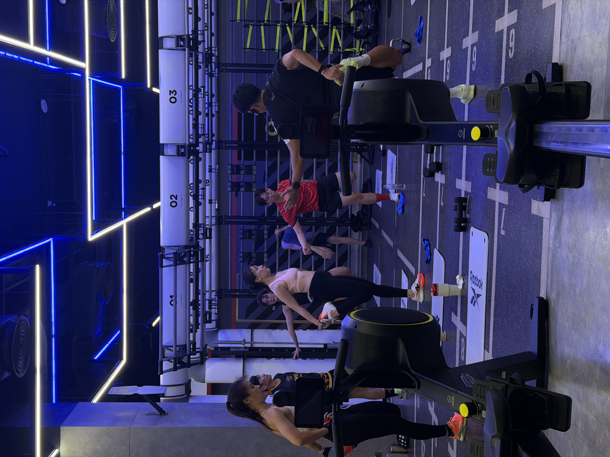 The image shows a group of people exercising in a gym. There are at least seven individuals visible, some of whom appear to be stretching. The environment is a modern fitness studio with blue and white LED lighting. Equipment such as rowing machines and weight racks are present. The flooring is a dark, textured surface with white markings. The gym appears to be well-lit and organized. The specific location of Nong Pa Khrang, Thailand, is not discernible from the visual information provided in the image.