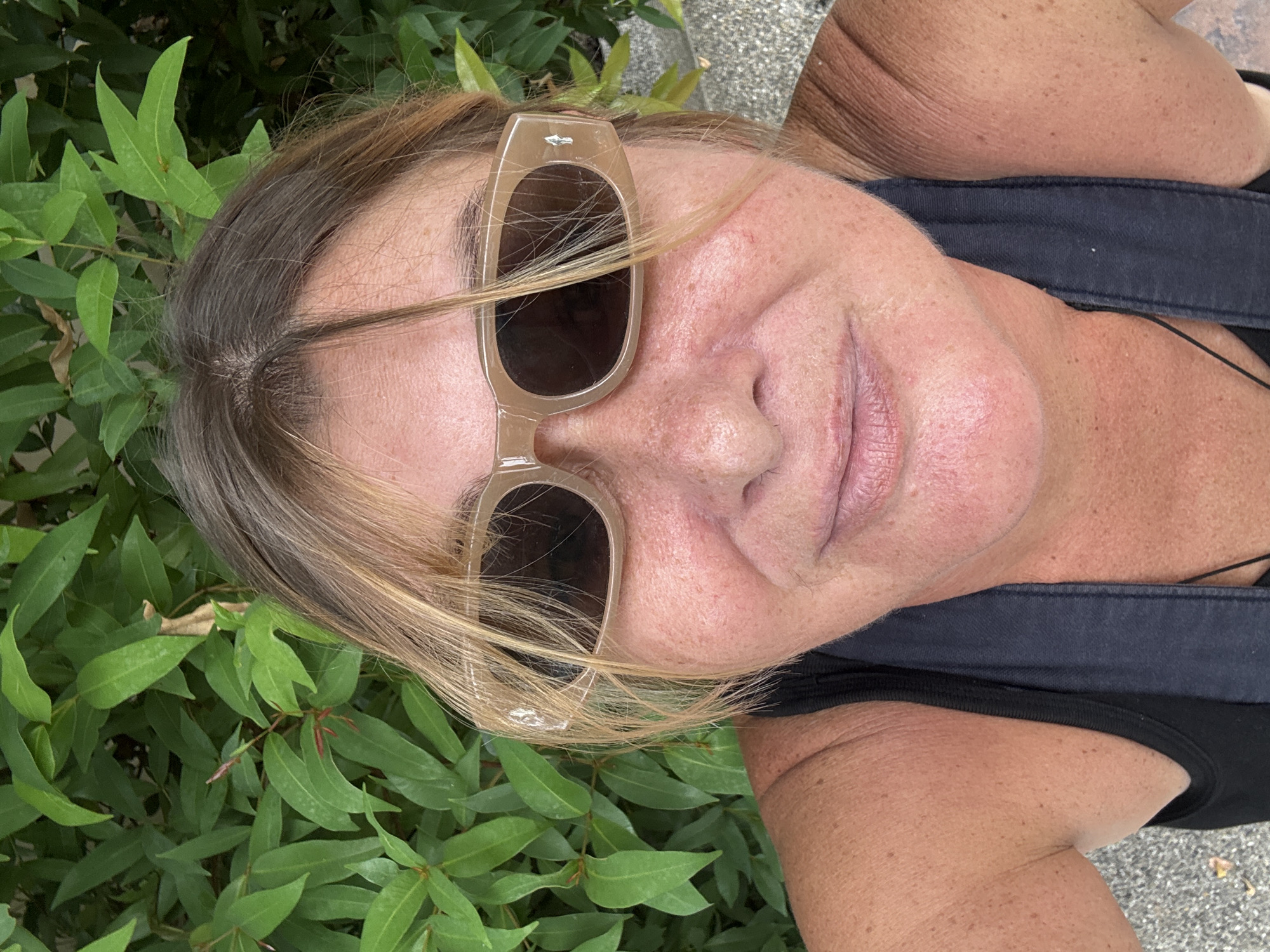 A selfie of a woman with blonde hair and sunglasses is taken outdoors in front of a green leafy bush. The woman is wearing a black tank top. The background shows lush green foliage, and part of a grey textured surface is visible in the lower right and left corners, suggesting an outdoor setting such as a park or garden.