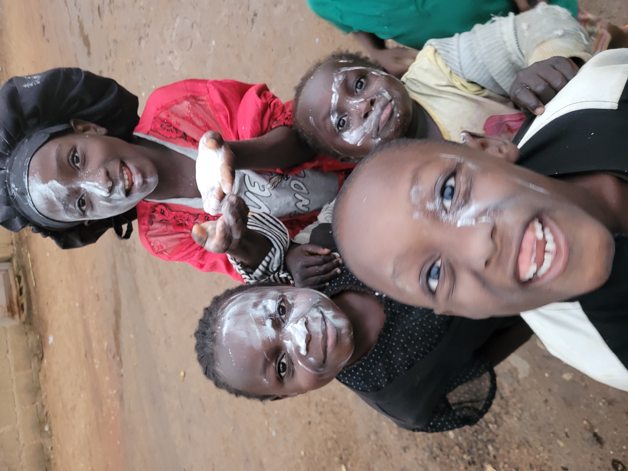In Bukuru, Nigeria, a group of young children are gathered, their faces and hands smeared with white powder, likely chalk or flour. The foremost child, with a wide, open-mouthed smile, is positioned in the foreground, their face taking up a significant portion of the frame. Behind them, several other children are visible, some looking directly at the camera, others with their faces partially obscured. One child, at the top of the frame, is wearing a black headwrap and a red garment, holding out their hands which are also covered in the white powder. The environment appears to be an outdoor, unpaved area, with loose dirt and debris on the ground. The overall impression is one of playful activity and shared experience among the children.
