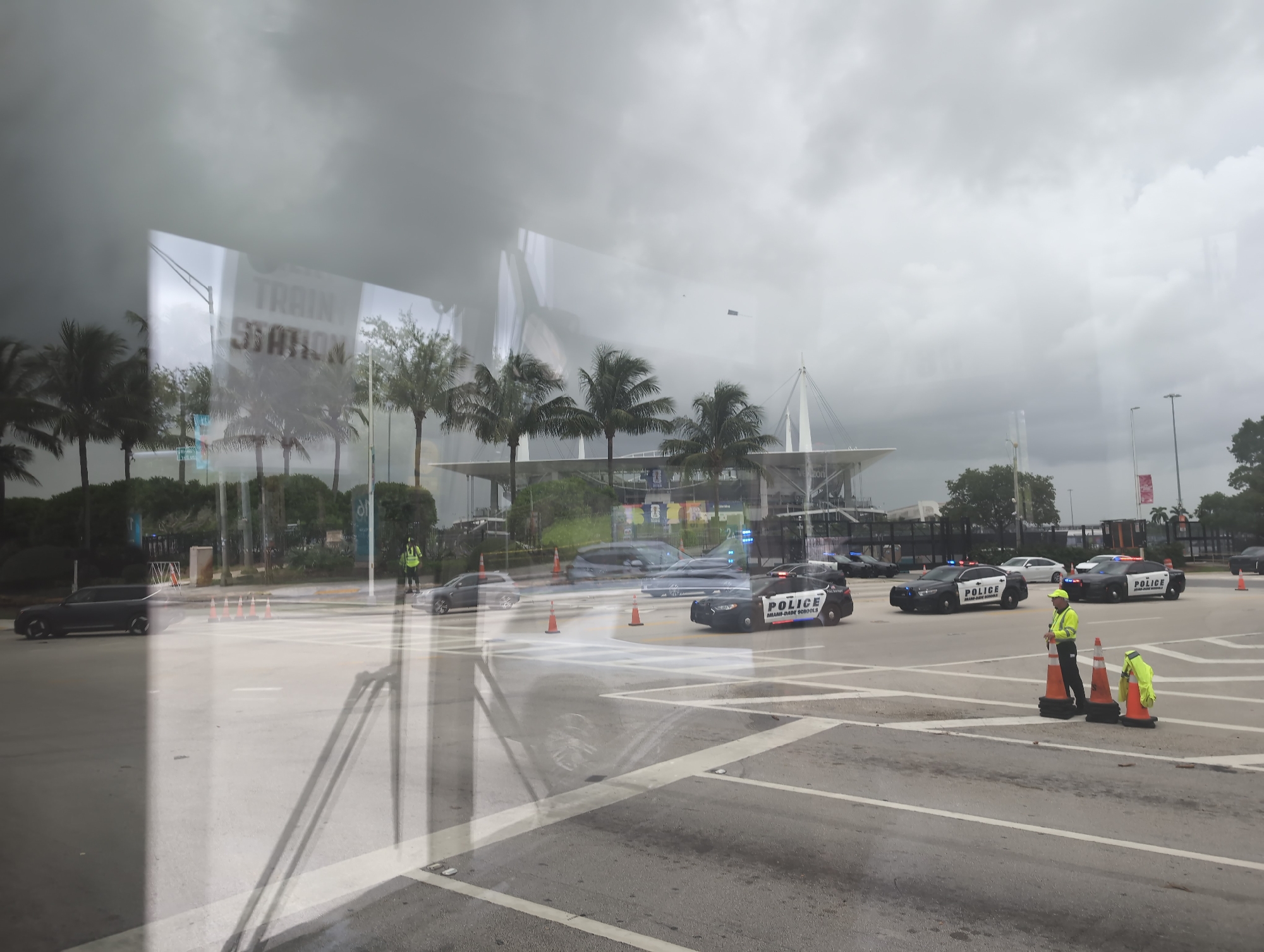 This image captures a scene outside a stadium in Miami Gardens, Florida, on a cloudy day. Reflections from a window obscure parts of the view. Several police vehicles, including marked cars with "POLICE MIAMI-DADE SCHOOLS" visible, are present. Two individuals in high-visibility yellow vests appear to be directing traffic or managing the scene. Orange traffic cones are strategically placed on the roadway. The stadium's distinctive architecture, with its white tent-like roof structure, is visible in the background, along with palm trees and other vegetation. The overall impression is of a controlled area, possibly related to an event or a security operation.