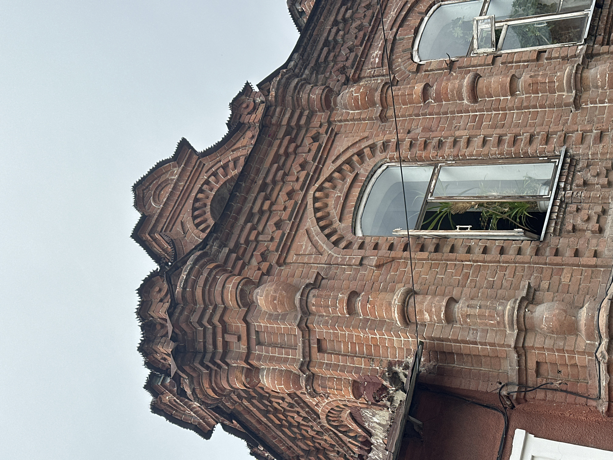 The image shows a close-up, low-angle view of the upper corner of an old brick building in Biysk, Russia, under an overcast sky. The building features ornate brickwork with decorative elements such as cornices, pilasters, and arched window frames. One large arched window is open, revealing houseplants inside. Another smaller arched window is partially open. Electrical wires are visible running along the facade. The brickwork appears weathered and in some areas, chipped or damaged, particularly along the roofline.