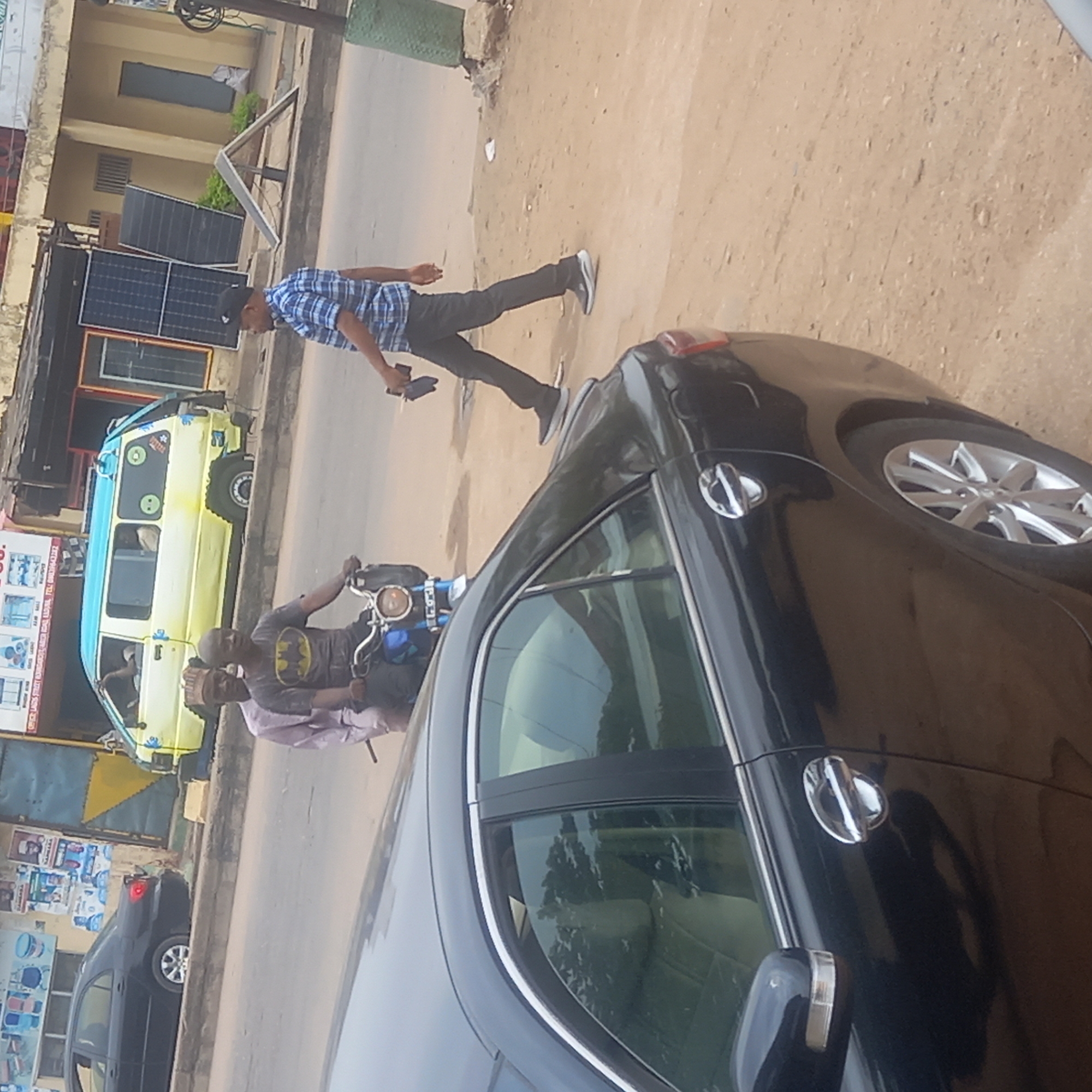 The image captures a street scene in Dadi Riba, Nigeria. A black car is prominently positioned in the foreground, partially obscuring the view. Further down the street, a yellow and blue van is parked. Two men are riding a motorcycle, with one wearing a Batman T-shirt. A man in a blue checkered shirt walks on the sidewalk. Buildings with solar panels are visible on the left side of the street. Various signs and posters adorn the storefronts.