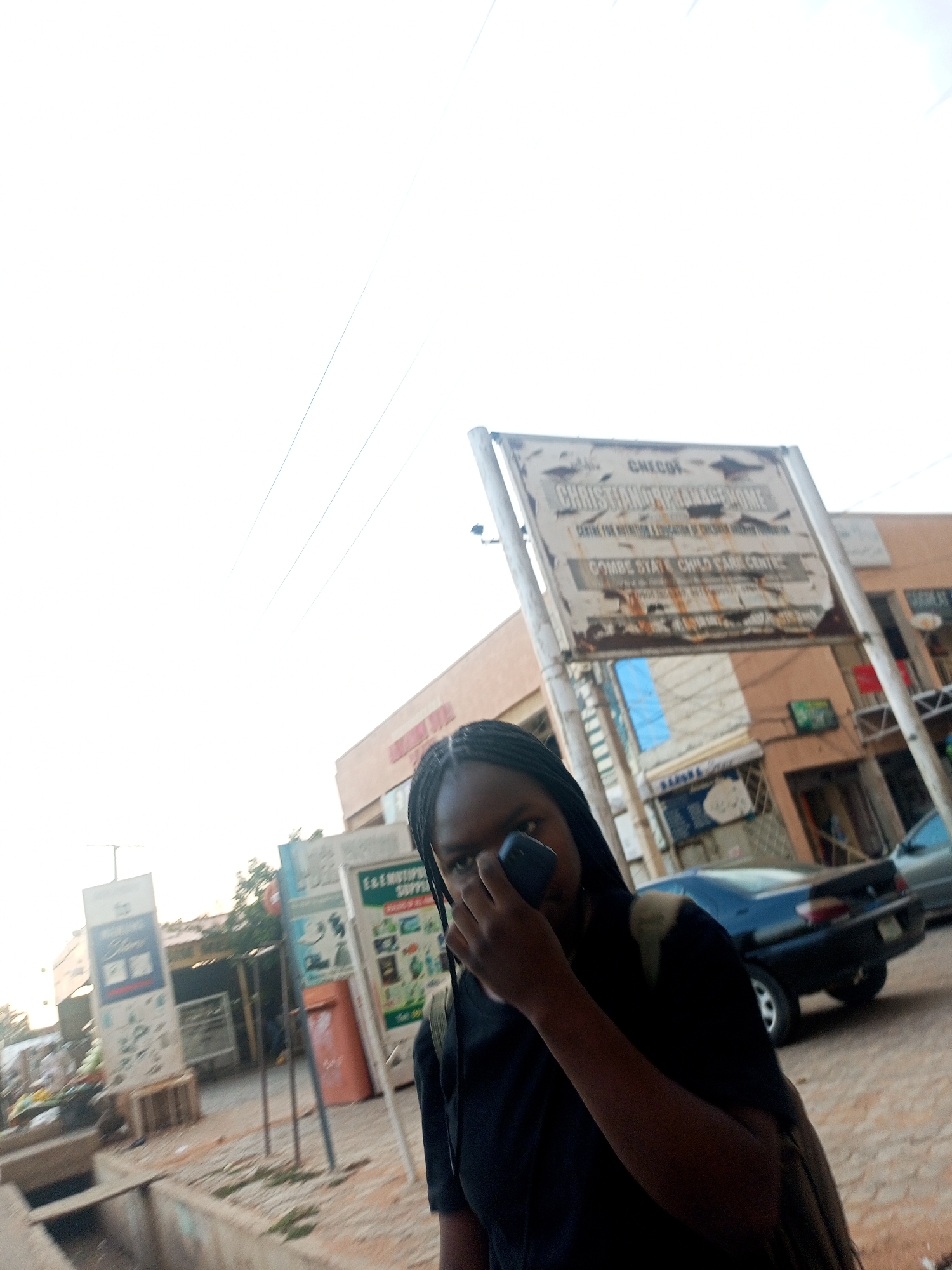 A woman holds a smartphone up to her face, standing on a sidewalk in Gombe, Nigeria. Behind her, a large sign reads "CHECKOK CHRISTIAN ORPHANAGE HOME, CENTRE FOR NUTRITION & EDUCATION OF CHILDREN WITH DISABILITIES, GOMBE STATE CHILD CARE CENTRE". Shops and vehicles are visible along the street, including a dark blue sedan. The sky is overcast.