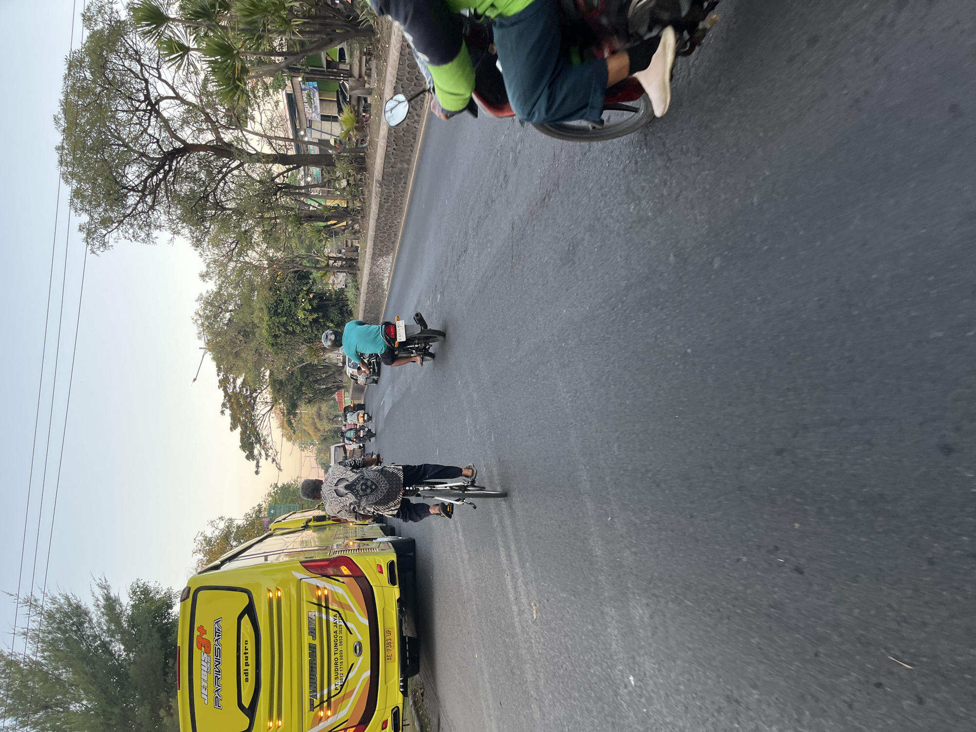 This is an outdoor, eye-level shot of a road with multiple vehicles and pedestrians. The scene appears to be in Ngaban, Indonesia, given the context. In the foreground, a person rides a motorcycle, wearing a green long-sleeved shirt, dark pants, and light-colored shoes. To the left, a person on a bicycle is moving away from the camera, wearing a patterned shirt and dark pants. Further ahead, a yellow bus with "JETBUS 3+" and "PARIWISATA" written on it is stopped in traffic. Several other motorcycles are also visible on the road. The environment is urban, with trees lining the side of the road and buildings in the background. The sky is clear and light, suggesting it is either early morning or late afternoon. The road surface is asphalt, and there is a stone wall bordering the right side of the road.