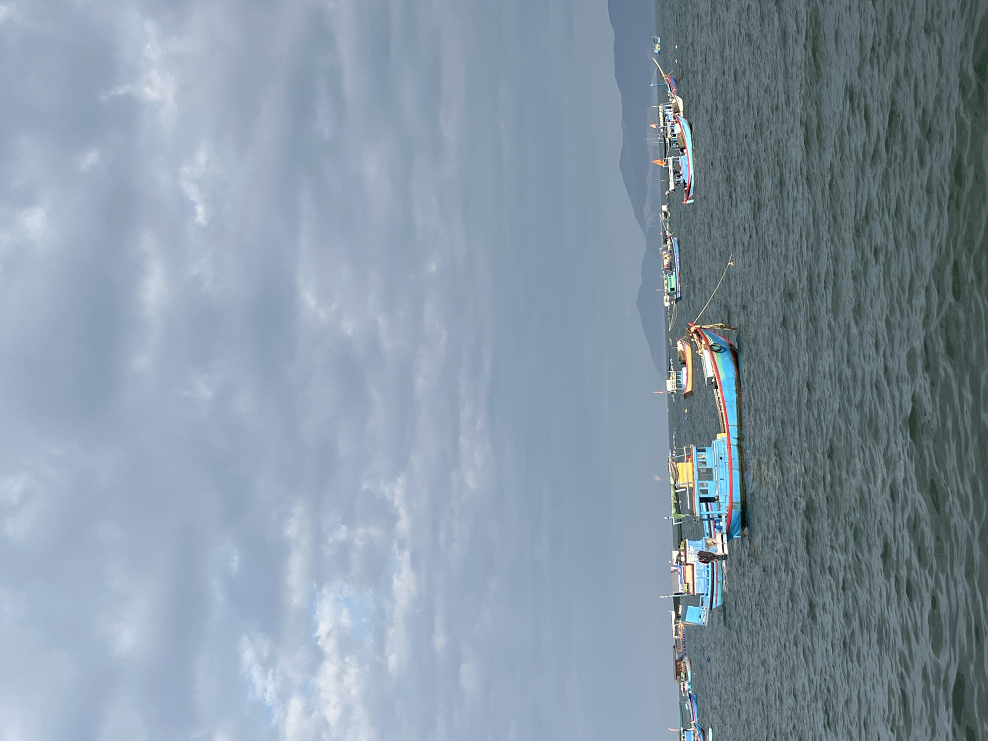 The image captures a maritime scene in Bac Nha Trang Ward, Vietnam, under a uniformly overcast sky. The upper half of the frame is dominated by a dense layer of light to medium grey clouds, with minimal variation in texture. Below the cloudy expanse, a distant, hazy line of dark, undulating mountains or hills extends across the horizon, separating the sky from the water. The lower half of the image consists of choppy, dark blue-grey water, showing consistent surface ripples and small waves.
Numerous small to medium-sized fishing boats are scattered across the water, appearing to be anchored or stationary. The boats are typically brightly colored; a prominent example in the mid-ground, slightly right of center, features a bright blue hull with a red lower stripe and a yellow-roofed cabin structure. A single individual wearing dark clothing is visible standing on the deck of this boat. Other boats, mostly of similar design with wooden hulls and small structures on deck, are visible in various positions, some featuring blue, red, green, or yellow accents. One boat in the immediate foreground on the left side, partially cropped, displays a red hull bottom and a light blue upper section, with a thick rope extending from its bow. The overall scene suggests a busy fishing area on a cloudy day.