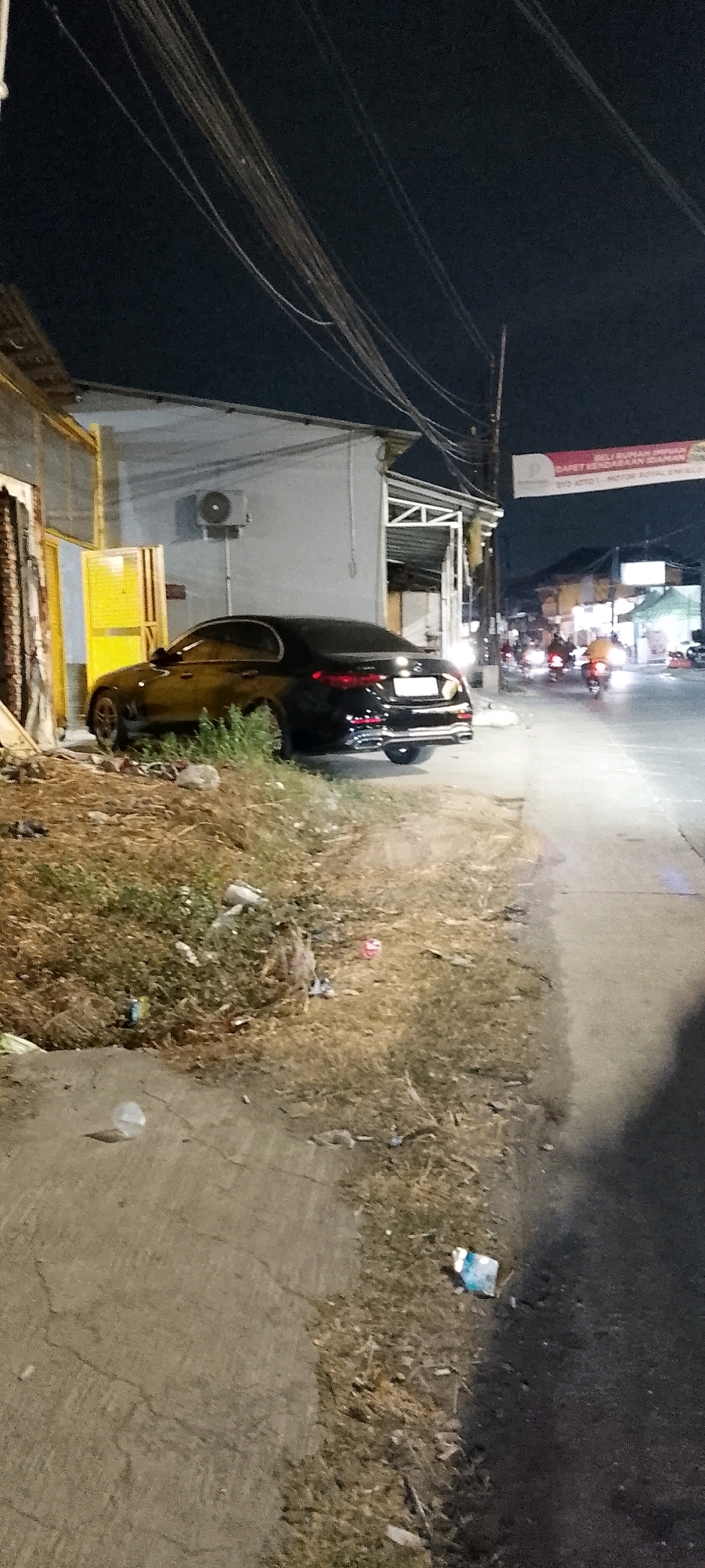 The image depicts an outdoor scene at night in the city of Satriamekar, Indonesia. A black car is parked on the side of a road, adjacent to a building with a yellow door and an open doorway. The building appears to be a shop or workshop. In the background, other vehicles, including motorcycles, are visible on the road, with their headlights illuminating the scene. The ground is covered with dry grass and scattered litter. Overhead, numerous power lines are visible against the dark night sky. A banner with text in Indonesian is also hung across the street. The overall impression is a dimly lit, urban roadside environment.