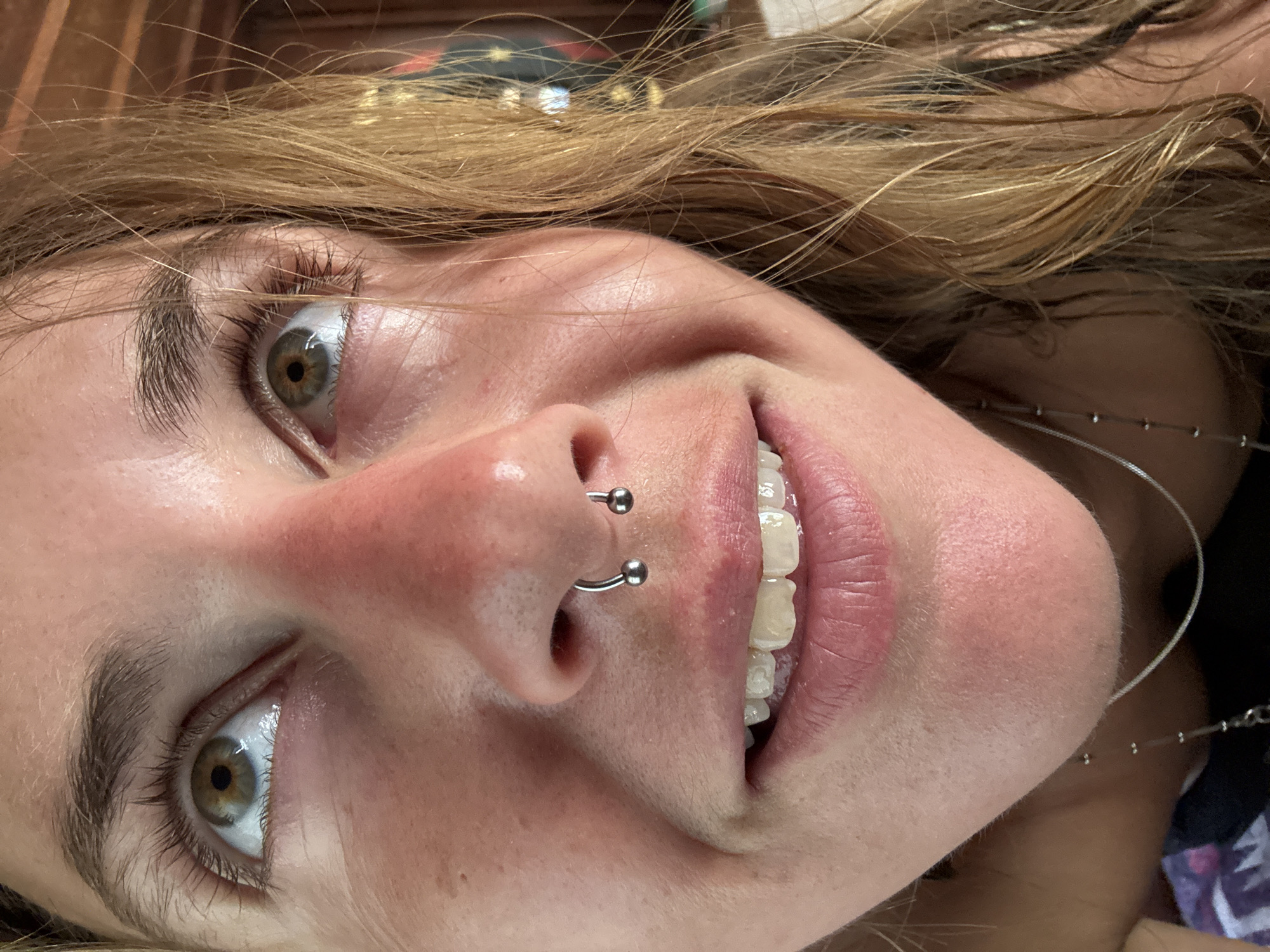 A close-up, low-angle, eye-level shot shows the face of a person with light brown, wet hair. The person has fair skin with some freckles and redness on the cheeks and nose. Their eyes are open and looking upwards. One eye is in focus, revealing a green-brown iris. The person has a septum piercing with a silver horseshoe-shaped ring. Their mouth is open in a smile, showing teeth and pink lips. A delicate silver necklace is visible around their neck. The background is out of focus, showing hints of a wooden structure and some lights. The image appears to have been taken indoors in natural light.