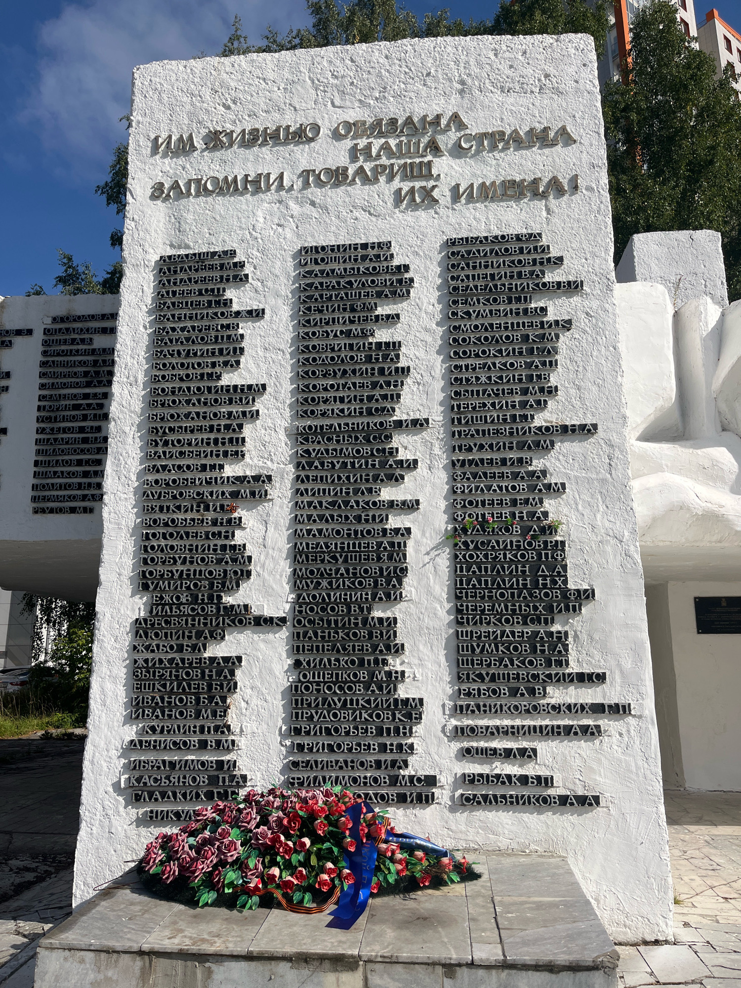 The image shows a white monument with golden lettering and a wreath of red roses at its base. The monument is located in the city of Perm, Russia, specifically in the Victory Park (Park Pobedy). The inscription on the monument reads "ИМ ЖИЗНЬЮ ОБЯЗАНА НАША СТРАНА ЗАПОМНИТЕ ТОВАРИЩИ ИХ ИМЕНА!" which translates to "Our country is indebted to them for their lives, remember, comrades, their names!". The monument lists the names of fallen soldiers in columns, with many of the names starting with Cyrillic letters such as "А", "Б", "В", "Г", etc. The background shows trees, a blue sky, and a modern building. The wreath at the base of the monument consists of numerous red roses with some green foliage and a blue ribbon. The overall scene is somber and commemorative, suggesting a memorial to soldiers who died during wartime.