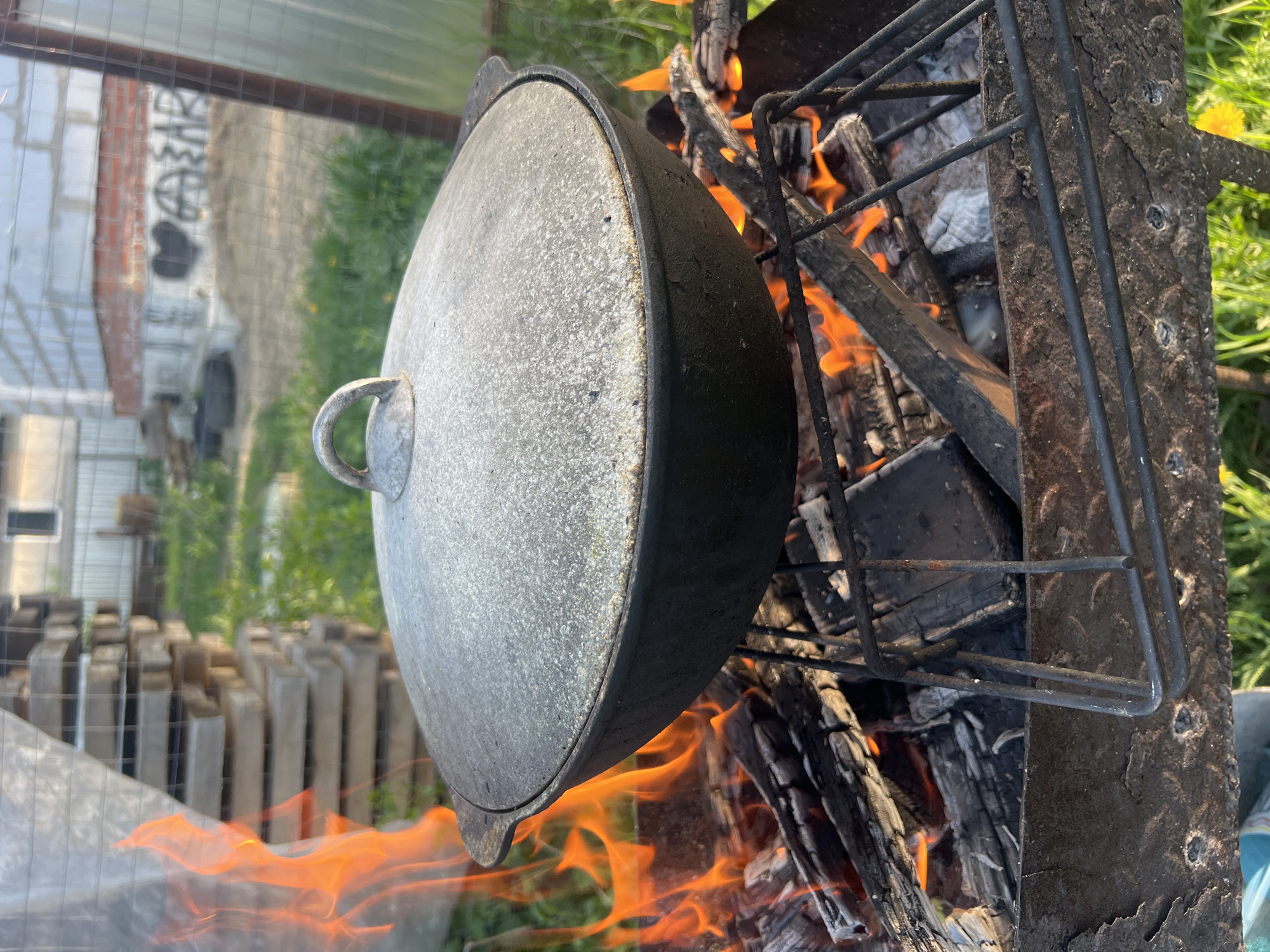 The image shows a cooking pot resting on a metal grate over a wood fire. The pot is large, round, and silver-gray with a looped handle on the lid. Flames are visible beneath and around the pot, licking up the sides. The grate is dark metal and appears well-used. In the background, out of focus, there are stacked wooden pallets and a fence or structure with a brick wall and what appears to be graffiti. Based on the location provided (Chaikovsky, Russia), the image likely depicts outdoor cooking in a rural or suburban setting.