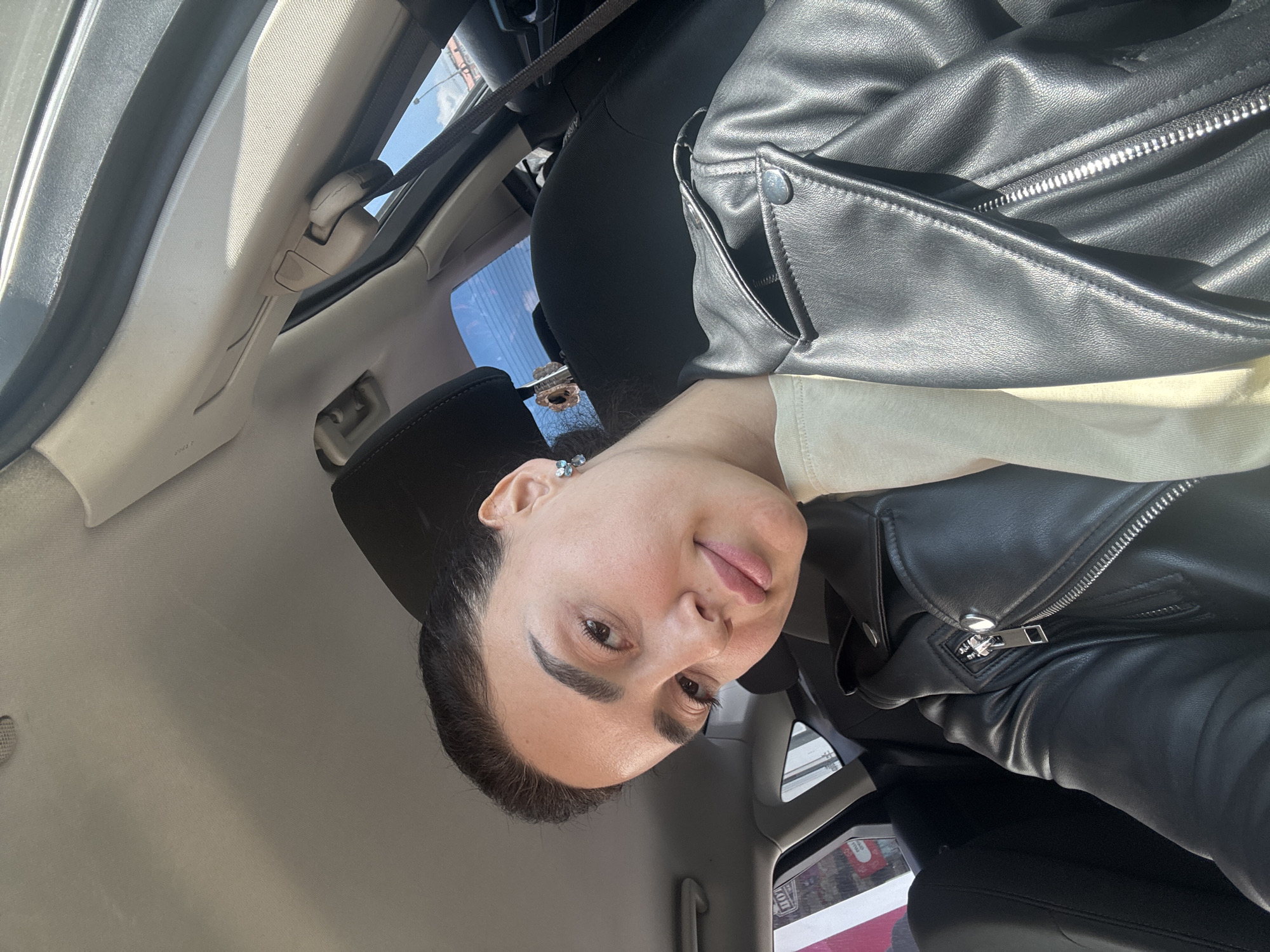 A woman is sitting in the driver's seat of a car. She's wearing a black leather jacket and a light-colored shirt. The car's interior is visible, including the headrest, seatbelt, and interior paneling. Outside the car window, there are buildings visible. Based on the urban environment and architectural style, the location is likely Tbilisi, Georgia.