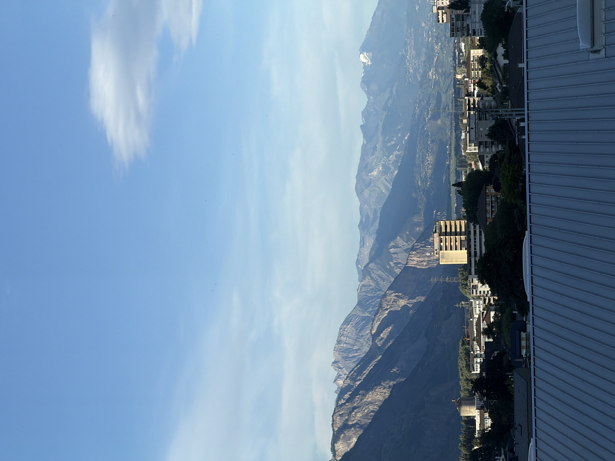 The image captures a panoramic view of the city of Martigny, Switzerland, with the Alps forming a dramatic backdrop. In the foreground, a corrugated metal roof dominates the bottom of the frame, suggesting a rooftop perspective. Beyond the roof, the cityscape unfolds, characterized by a cluster of buildings of varying heights and architectural styles. Several apartment buildings and houses are visible, interspersed with trees and greenery, indicating a residential area. One prominent multi-story building, possibly an office or residential complex, stands out with its beige facade and multiple windows. A large, cylindrical structure, resembling a silo or water tower, is also visible in the middle ground. Further in the distance, the majestic peaks of the Alps rise, their rocky slopes illuminated by the sun. Some of the higher peaks appear to be snow-capped, suggesting significant altitude. The sky is a clear blue, with a few wispy clouds scattered across it, hinting at good weather conditions. The overall scene depicts a harmonious blend of urban development and natural grandeur.