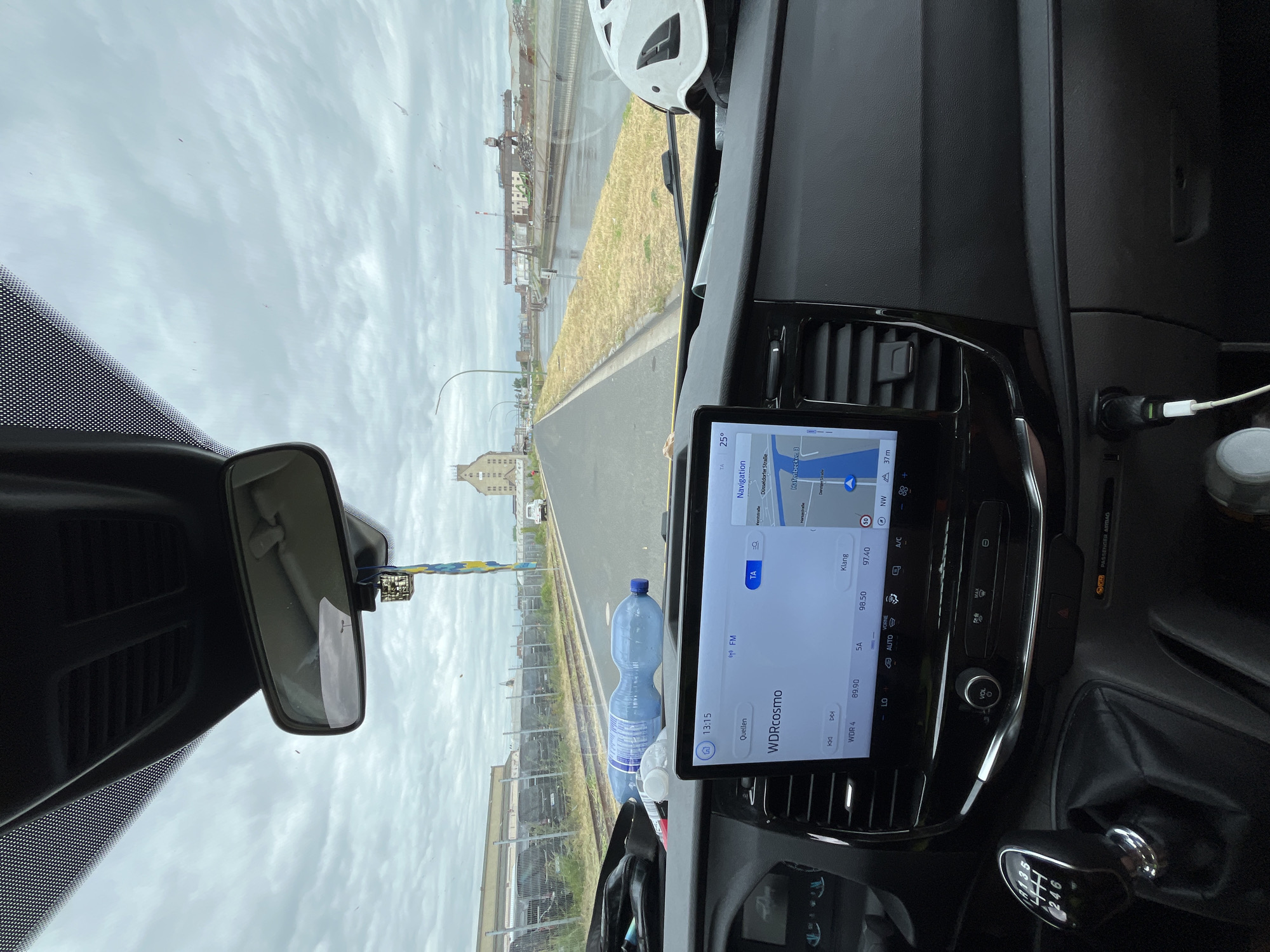 The image shows the interior of a vehicle, likely a van, looking forward through the windshield. The dashboard features a touchscreen infotainment system displaying navigation and radio information. The screen shows a map with the route highlighted, indicating a destination in Neuss, Germany. The radio is tuned to WDR Cosmo. A bottle of water is visible on the dashboard. Outside the vehicle, a road runs alongside a canal or river. In the distance, industrial buildings and cranes are visible under a cloudy sky. A white helmet is placed on the passenger side of the dashboard. The gear stick and part of the steering wheel are visible in the foreground.