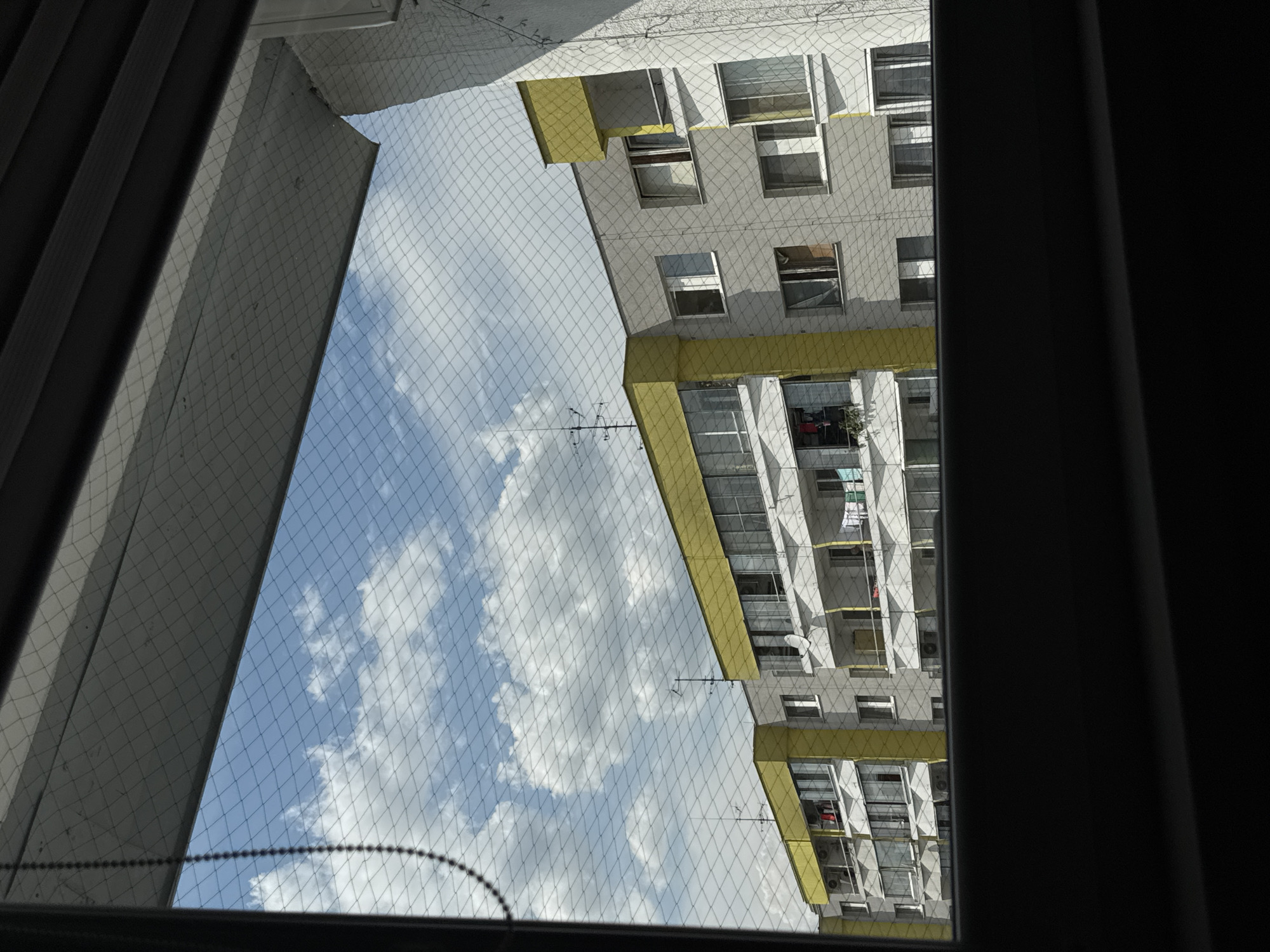 This is a photograph taken from an interior perspective looking out of a window. The window is covered by a fine mesh netting, which is visible throughout the image. Outside the window, a pale blue sky with scattered white clouds is visible. In the mid-ground, a multi-story residential building is seen. The building has a light gray facade with bright yellow accents on the upper floors and balconies. Many windows and balconies are visible, some with laundry hanging out to dry. The building appears to be an apartment block. The image is likely taken in Bratislava, Slovakia, as indicated by the architectural style and context. The foreground is dominated by the dark frame of the interior window.