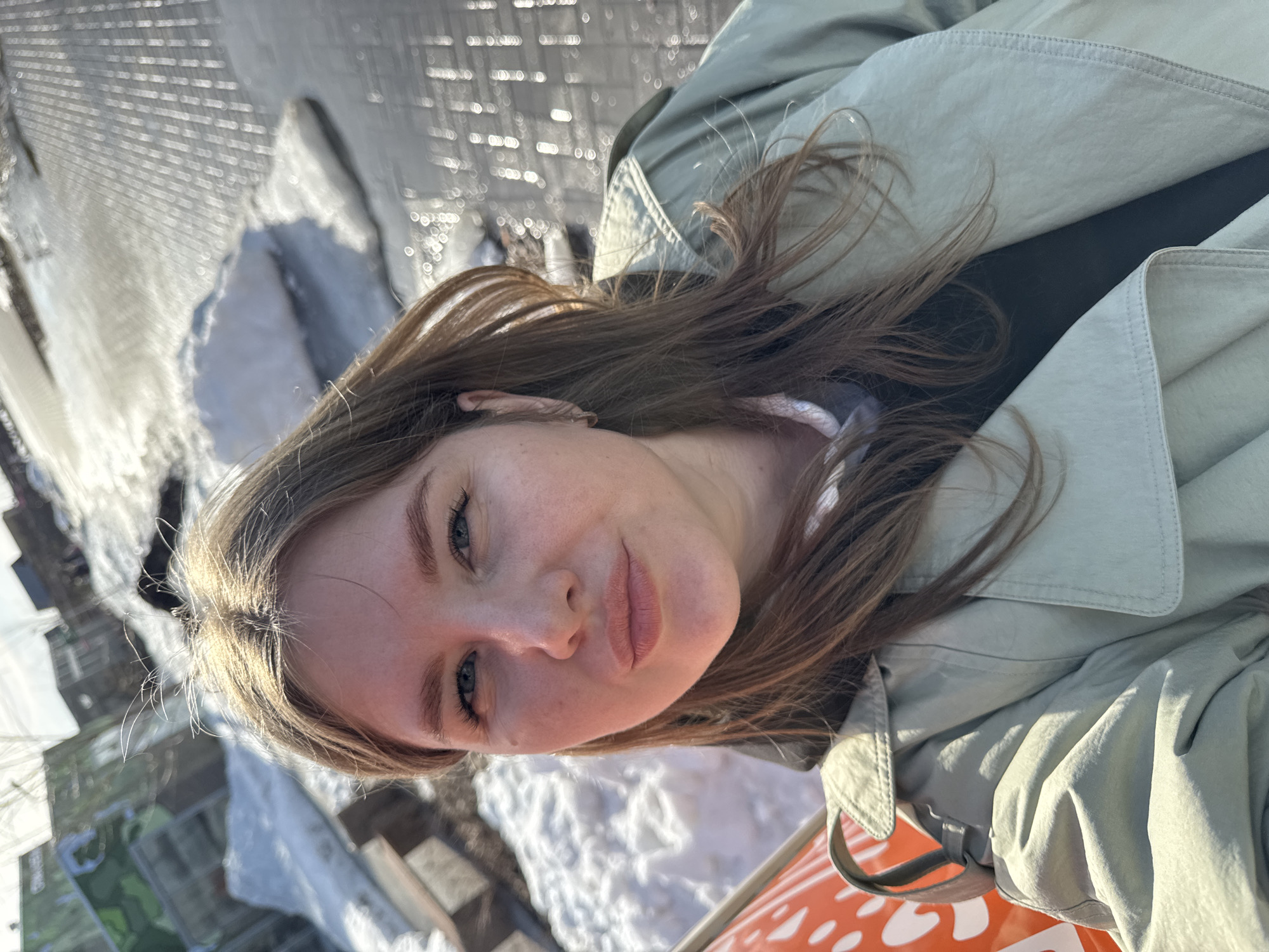 The image depicts a selfie taken by a fair-skinned female with light brown hair. She is wearing a light green trench coat. The background shows a snow-covered area, along with what appears to be a metallic, textured structure. To the right of the frame, part of an orange object with white spots is visible. The photograph was taken outdoors, seemingly in the city of Perm, Russia.