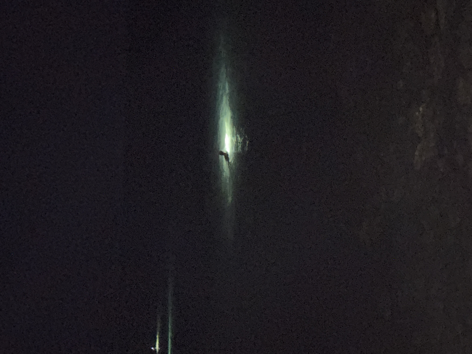 At night, a person is seen swimming in the dark water, illuminated by a bright light source. The water appears calm, with ripples visible around the swimmer. In the distance, another light source is visible, indicating a possible boat or other illumination. The background is dark and indistinct, suggesting an outdoor environment with minimal ambient light. The scene takes place in the sea waters of Agios Tychona Community, Cyprus.