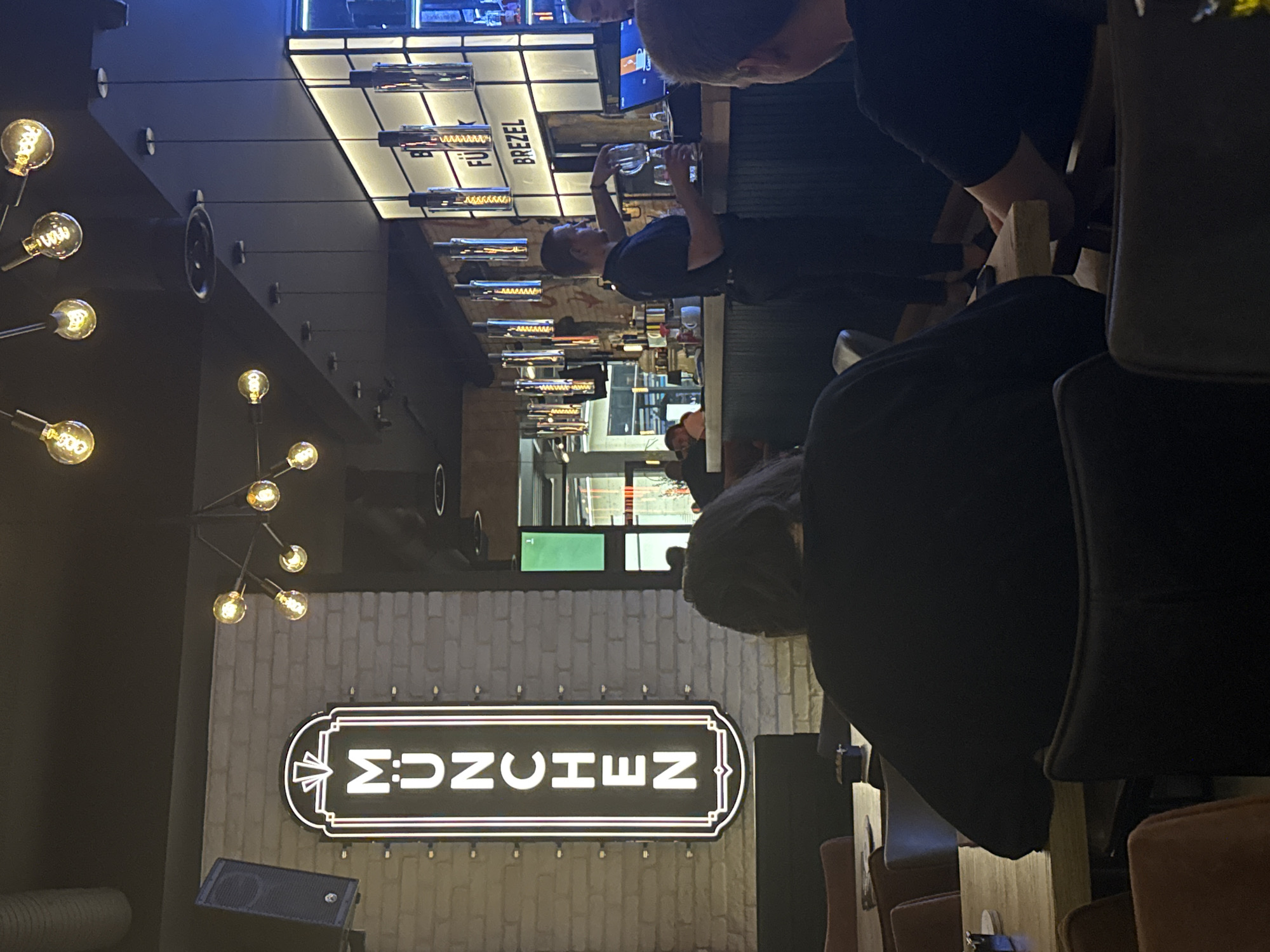The image depicts the interior of a dimly lit restaurant or bar. In the foreground, a person with dark, long hair wearing a black shirt is seated at a table, their back to the camera. To their right, another person, also with dark hair and wearing a black shirt, is visible from the back. The environment features exposed brick walls and industrial-style lighting fixtures hanging from the ceiling. A prominent neon sign spelling out "MÜNCHEN" in vertical, white lettering is mounted on the brick wall to the left. In the background, a bar area is visible with a bartender pouring a drink into a glass. Above the bar, a backlit sign displays the word "BREZEL." Several other patrons are seated at tables or standing around the bar area. The overall atmosphere suggests a casual dining or drinking establishment.
