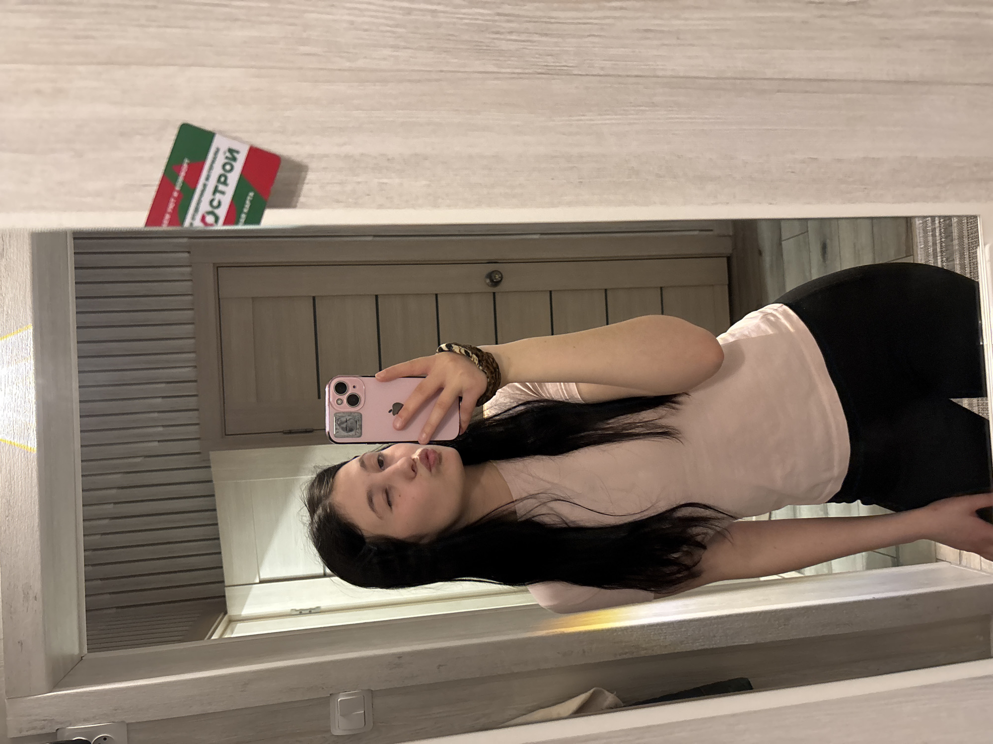 A young woman is taking a selfie in a mirror. She is wearing a light pink shirt and black pants. She is holding a pink iPhone in her left hand and puckering her lips. Her hair is long, dark, and straight. There is a discount card on top of the cabinet with the Russian word "Острой" on it. The location is Фоки, Russia.