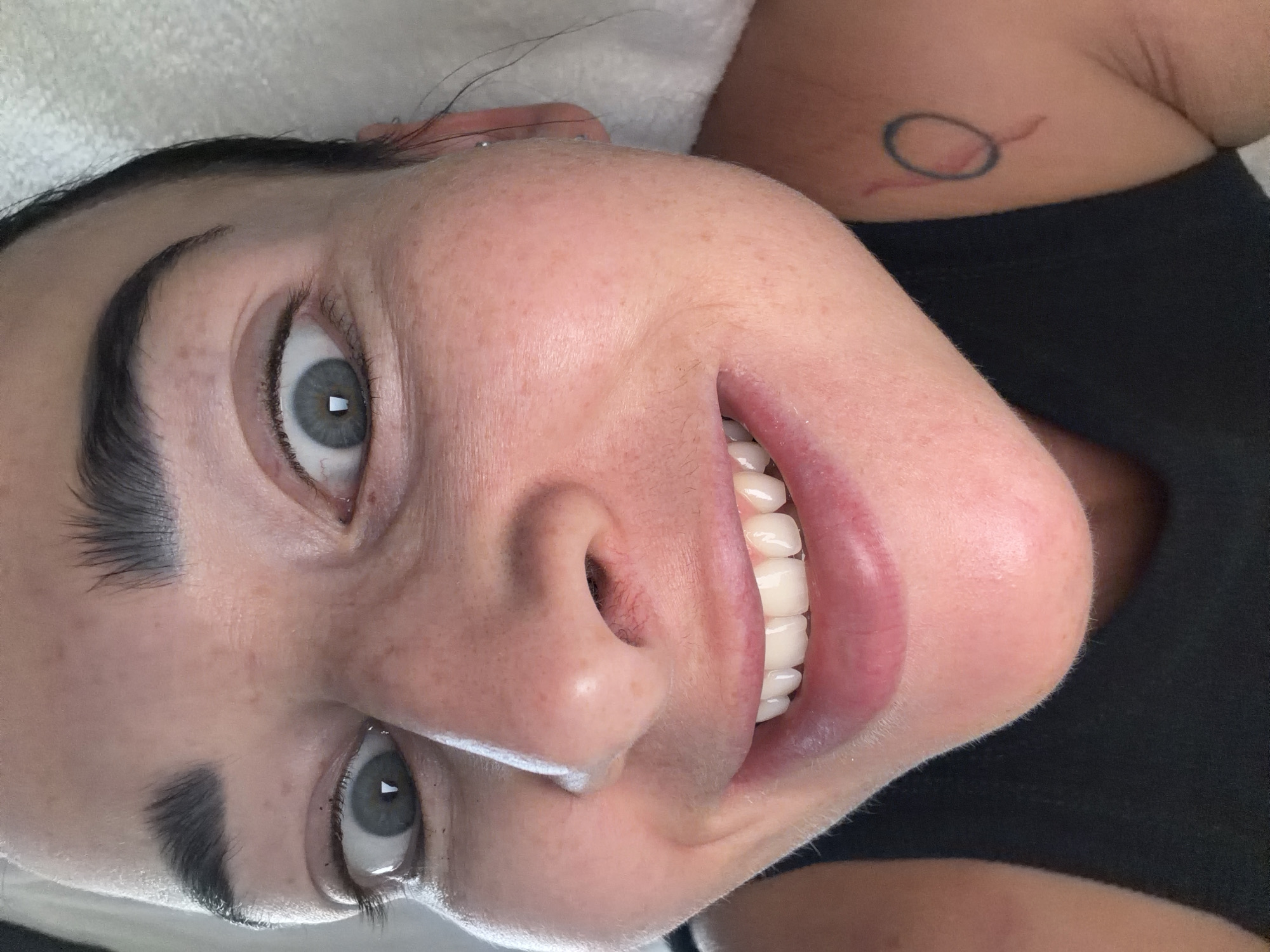 A close-up, rotated view of a Caucasian woman with pale skin, freckles, and dark hair. She has arched eyebrows and light-colored eyes with dark eyeliner. She is smiling, revealing white teeth. Part of her nose and chin are visible. A tattoo of a circle with a tail-like extension is visible on her shoulder. She is wearing a black shirt. The background includes a white or light-colored surface. There is no evidence of the image being in Saint Petersburg, Russia.