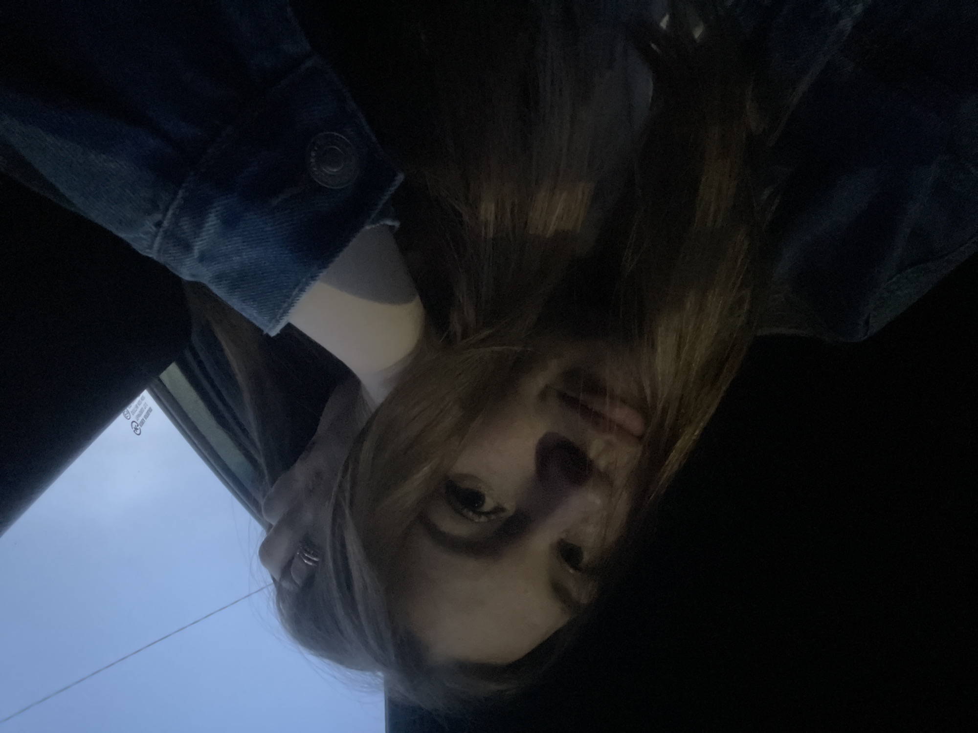 The image shows a woman with long, light brown hair, wearing a denim jacket. She is looking down at the camera, holding her head with a hand adorned with a ring. The background includes a car window and what appears to be a glimpse of the sky. The lighting is dim, with shadows covering the bottom right portion of the frame. No information indicates the city of Chaikovsky, Russia.