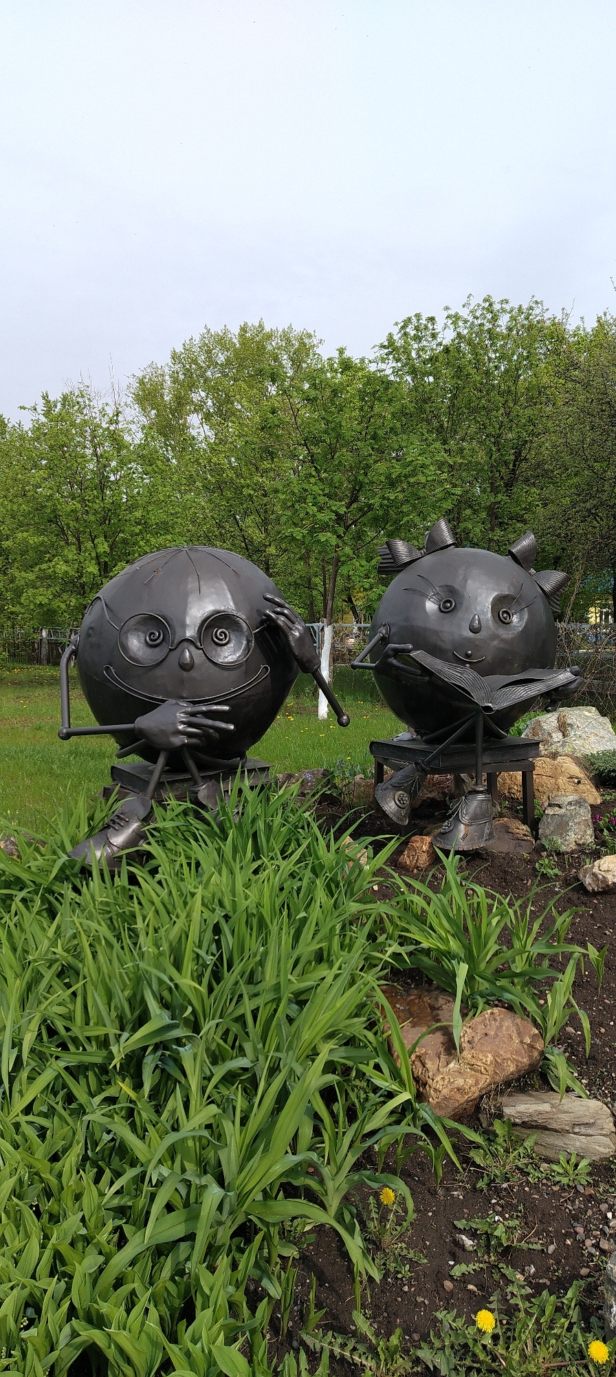 The image shows two metal sculptures of anthropomorphic round figures in a park-like setting, likely in Chaikovsky, Russia. The figure on the left wears glasses and is touching the frame with one hand, while the other hand rests on their lap. The figure on the right is reading a book with a pair of decorative bows adorning its head. Both figures sit on small stools and are positioned within a flowerbed overgrown with tall green plants. The background includes a grassy area and trees with green foliage. The sky appears overcast.