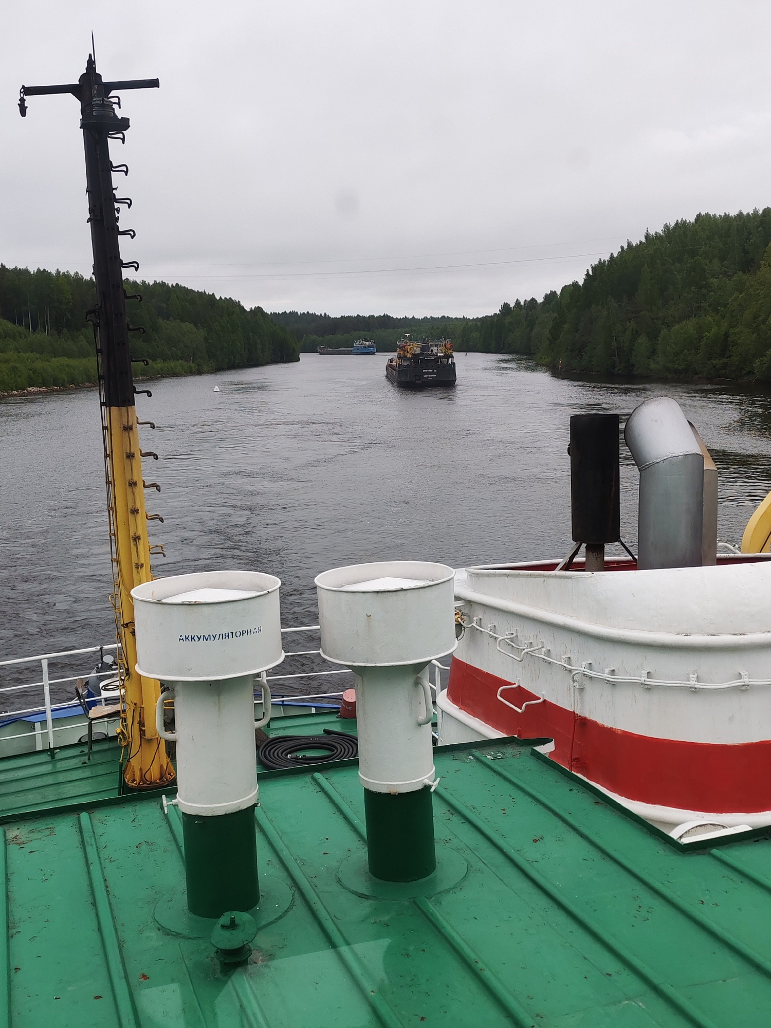 Captured from the deck of a vessel in Depo, Russia, the scene overlooks a waterway. Two barges are visible on the water, with tree-lined banks on either side. The overcast sky suggests a cloudy day. The foreground reveals details of the vessel's deck, including ventilation shafts marked with Cyrillic script ("АККУМУЛЯТОРНАЯ"), and other nautical equipment such as a mast and exhaust stacks. The deck is painted in green, white and red, with additional features of ropes and tubes visible.