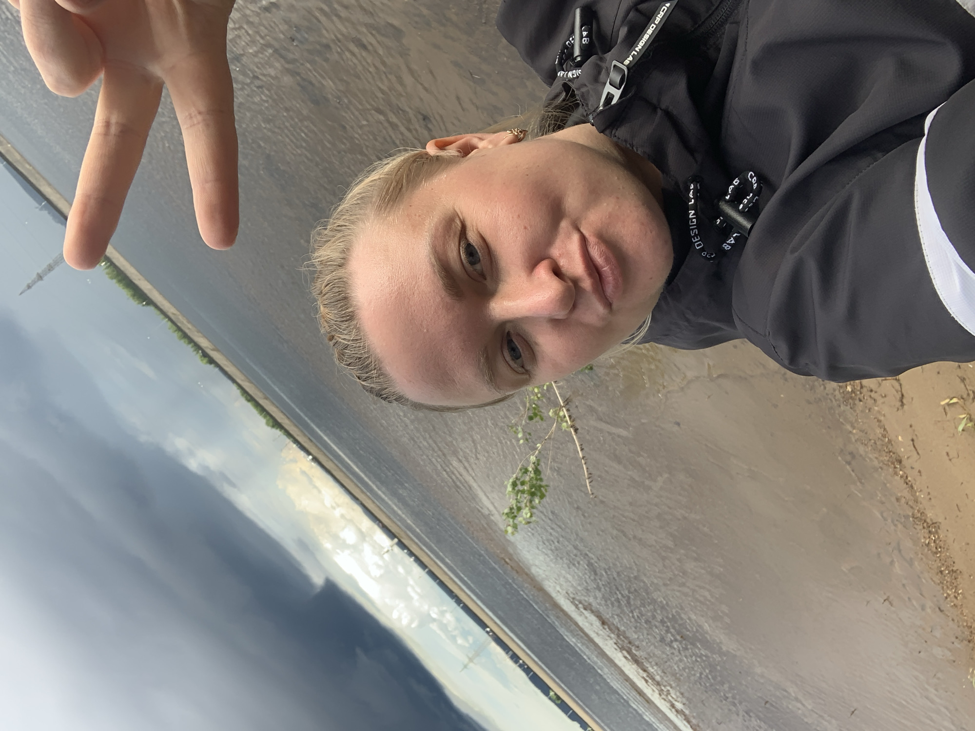 A close-up selfie captures a person, likely a young adult female, standing outdoors by a body of water. Her face, with blonde hair and fair skin, is angled slightly towards the camera, displaying a neutral expression with a subtle pout. She makes a peace sign gesture with her right hand, positioned in the upper left of the frame. She wears a black jacket with a white stripe on the right arm, and text reading "CRAP DESIGN LAB" is visible on the jacket's straps and drawstring.
The background features a wide body of water, possibly a river or large lake, with a narrow, light-colored shoreline to her right. The water's surface reflects the overcast sky, which shows expansive dark grey clouds dominating the distant horizon, suggesting a potentially stormy or moody atmosphere. A small branch with green leaves is visible near the water's edge in the foreground. In the far distance across the water, a faint landmass with some greenery and a tall, slender structure can be seen. The overall lighting indicates daytime. This scene is set near Noviy, Russia.