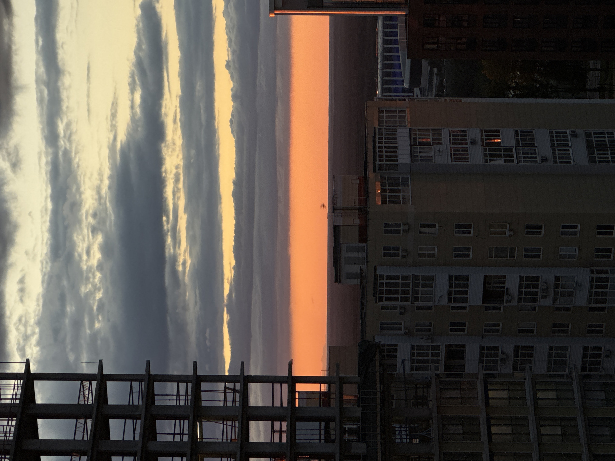 The image shows a cityscape at either sunrise or sunset. In the foreground are unfinished concrete building frames, partially obscuring the view. Adjacent to this is a tall residential building with rows of windows, some lit from within. Beyond the buildings, the sky is divided into three distinct sections: a section of heavy clouds, then a clear orange expanse, and finally a strip of lighter, sunlit clouds. The image is rotated ninety degrees counter-clockwise.