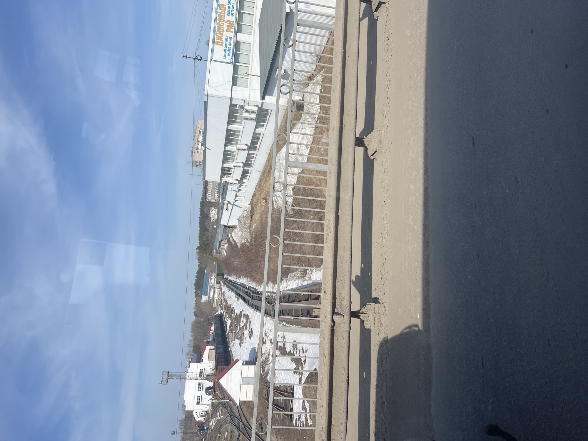 The image depicts a view from an elevated position, likely a bridge or upper floor, overlooking a railway yard. The scene includes railway tracks, trains, and buildings, with some snow visible on the ground. A building with the sign "ДЖИНСОВЫЙ РАЙ" (Jeans Paradise) in Russian is visible. The location is likely the city of Chaikovsky, Russia. There are no people visible in the image. The dominant features are the railway infrastructure and the surrounding buildings under a blue sky with some cloud cover.