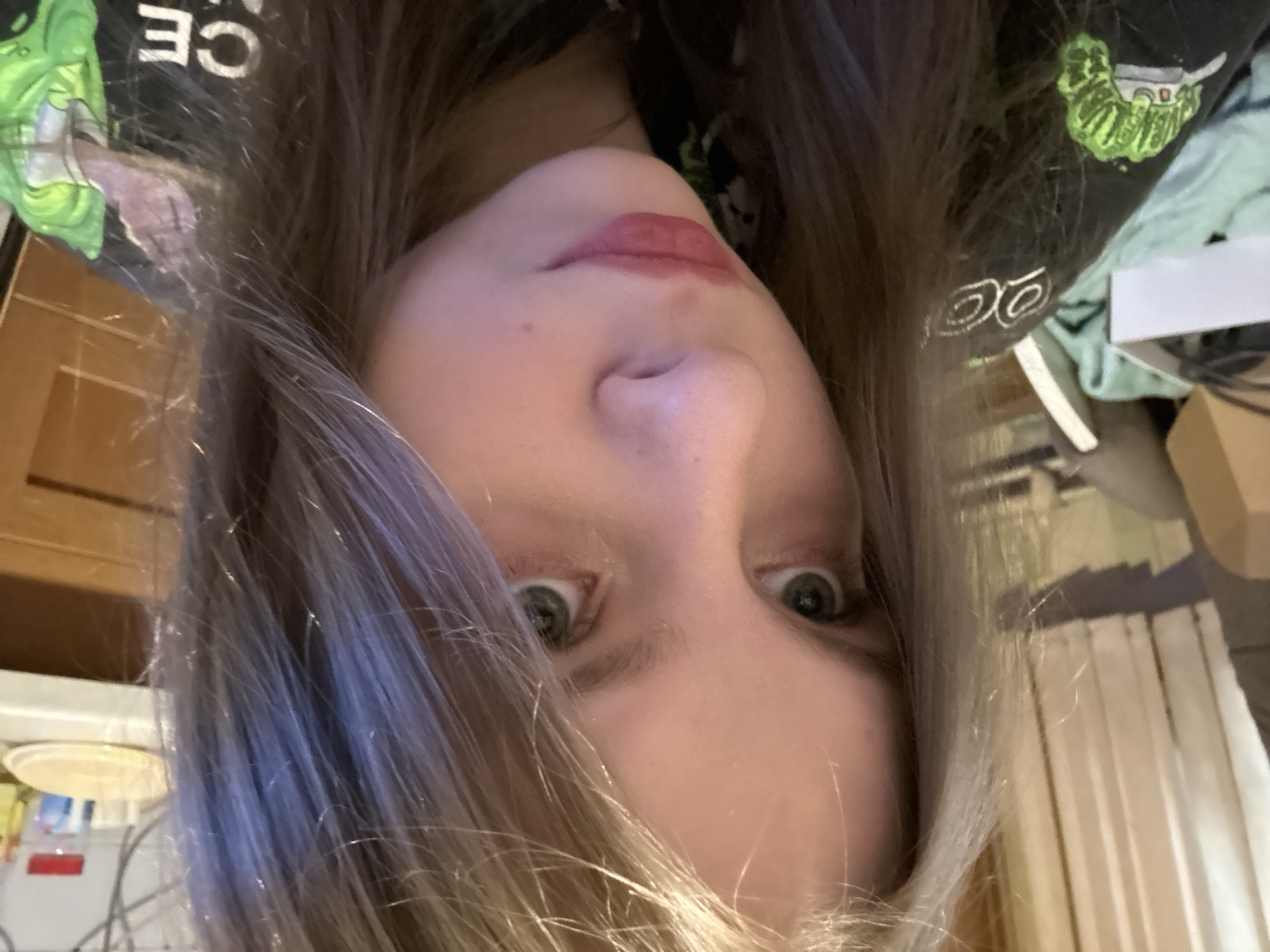 A close-up, upside-down selfie of a person with light skin, green eyes, and ash-blonde hair framing their face. They are wearing lipstick. A black graphic tee-shirt with neon green and white details is partially visible around the head. The background is cluttered and out of focus with glimpses of furniture and household objects. The location appears to be an indoor setting, potentially a home. There is no evidence to suggest that the image was taken in the city of Moscow, Russia.
