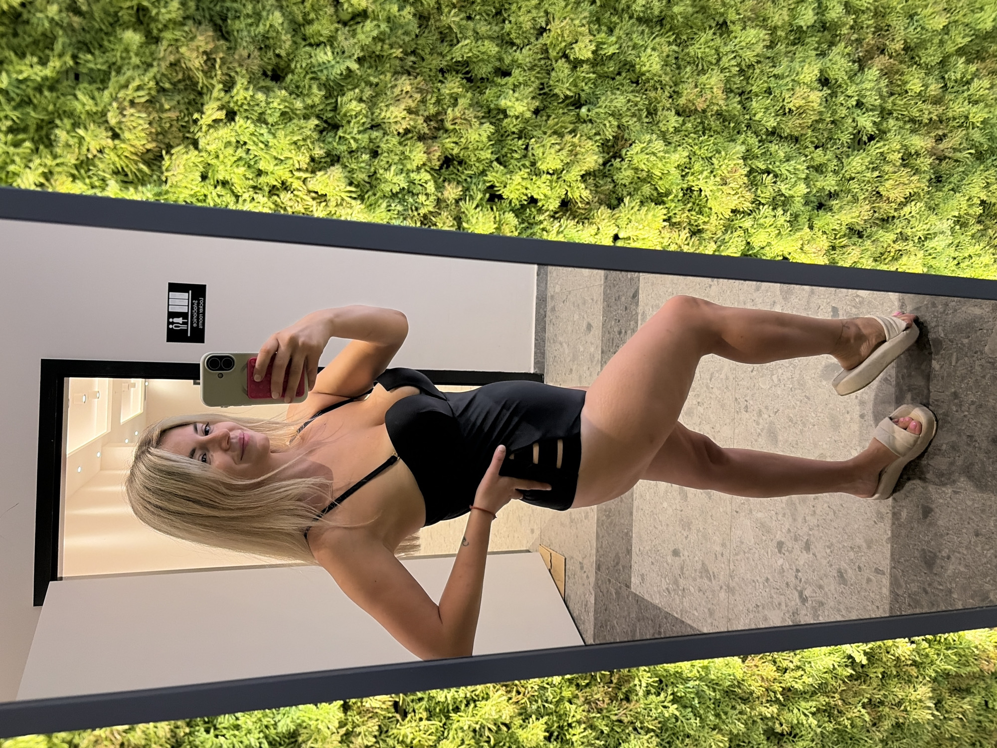 A woman with blonde hair, wearing a black swimsuit and white slides, takes a mirror selfie in a modern indoor space. She is standing, facing the mirror and holding a smartphone in her right hand. Her left hand is placed on her hip. The background of the mirror reflection shows a white wall with overhead lighting, a sign indicating "Locker Room," and a lush green moss wall to her right. The floor is tiled with grey and white patterns. The location is the exterior of a spa or wellness center in Podgorica, Montenegro.