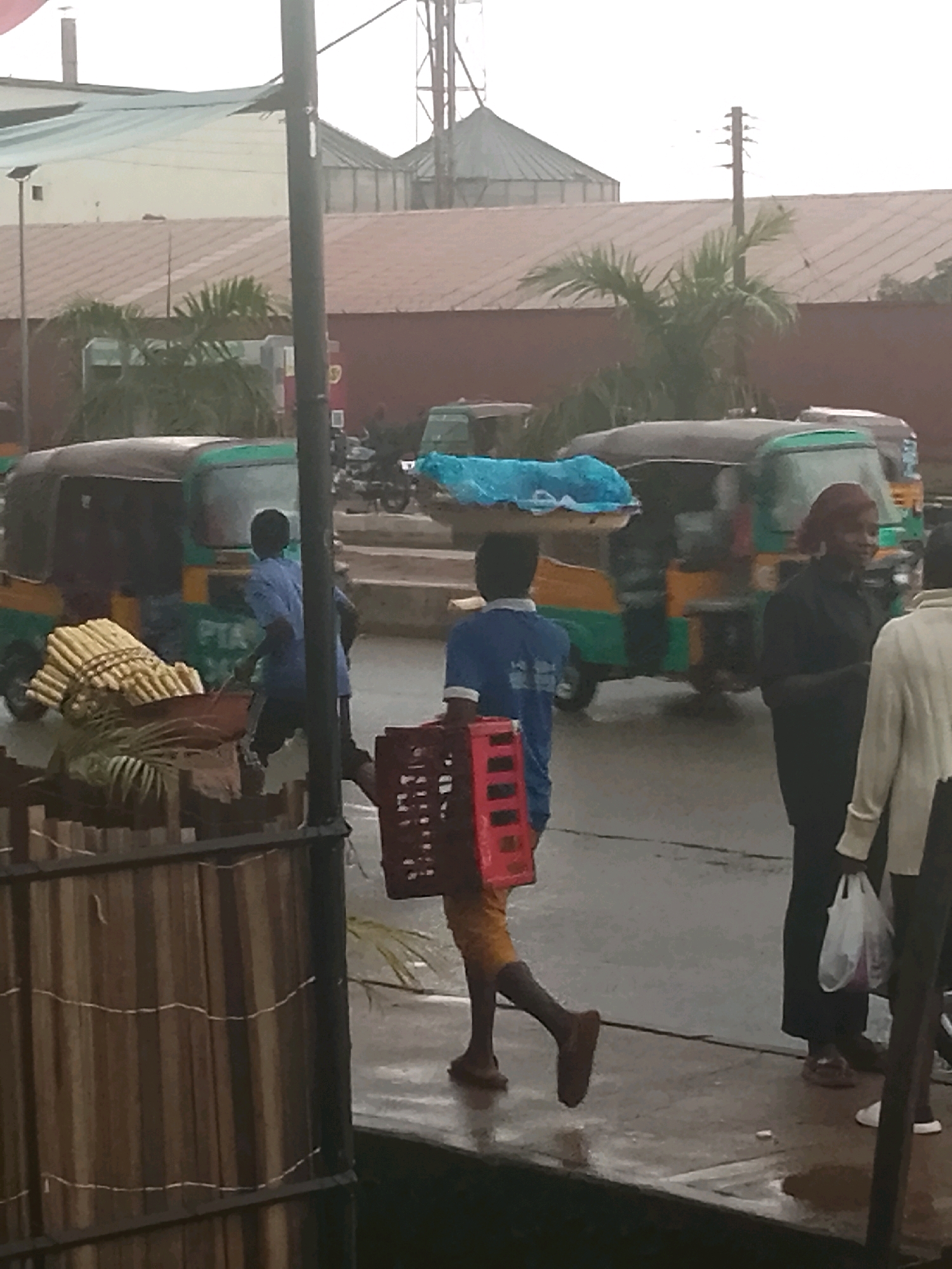 In Gura Top, Nigeria, on a wet street with reflective puddles, a young person in a blue t-shirt and yellow shorts walks from right to left, carrying a red plastic crate in their right hand and a bundle of sugarcane on their head. They are walking past a green and yellow auto-rickshaw, which is stationary. To the right of the auto-rickshaw, a woman in a black top and dark pants stands holding a white plastic bag. Further to the right, another person in a light-colored jacket and dark pants is partially visible. In the background, there are buildings with brown tiled roofs and palm trees. A tall pole with multiple horizontal extensions stands to the left of the center.