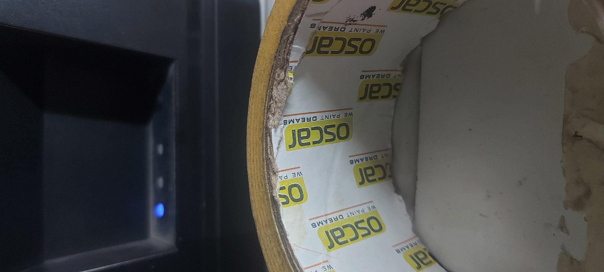 The image features a roll of "Oscar" brand masking tape positioned in front of a black electronic device with a blue light. The masking tape roll has a cardboard core printed with the brand name "Oscar" and the tagline "WE PAINT DREAMS" in yellow lettering. The tape itself is light brown. The core of the tape roll is partially empty and appears used. The black electronic device has a minimalist design with a rectangular shape and a glowing blue LED light. The setting appears to be indoors, based on the presence of the electronic device and the lighting conditions. The location information (Tashkent, Uzbekistan) is not evident from the image alone, as there are no clear contextual indicators.