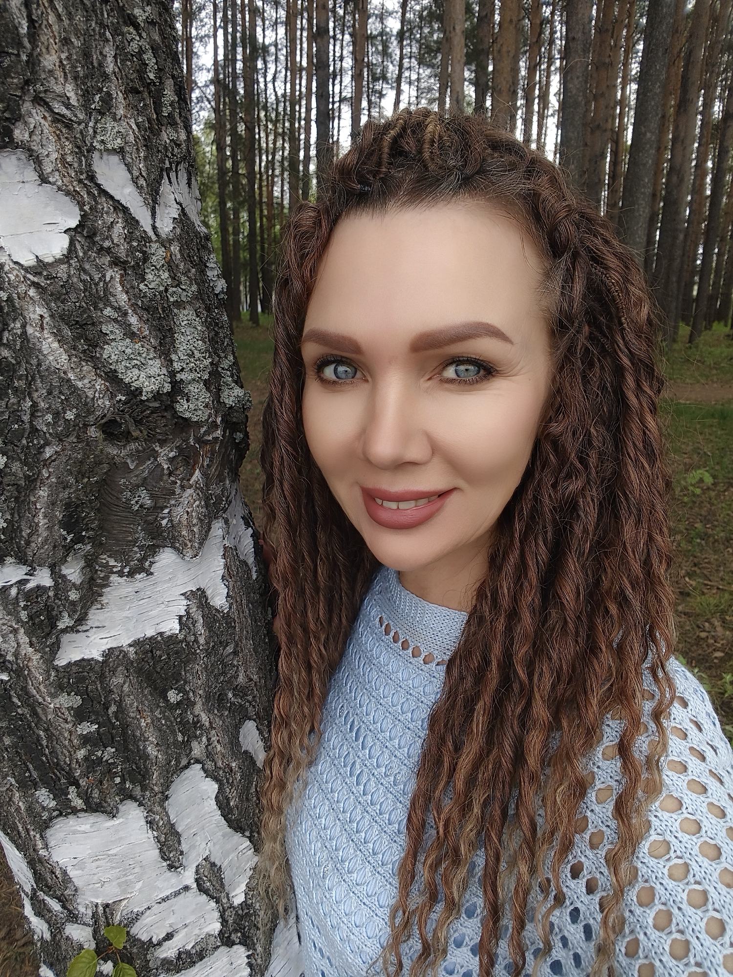 A fair-skinned woman with brown dreadlocks and light blue eyes poses next to a birch tree in a forest. The location is identified as the city of Unknown, Unknown. The woman is wearing a light blue, patterned sweater. The forest has many tall, thin trees in the background.