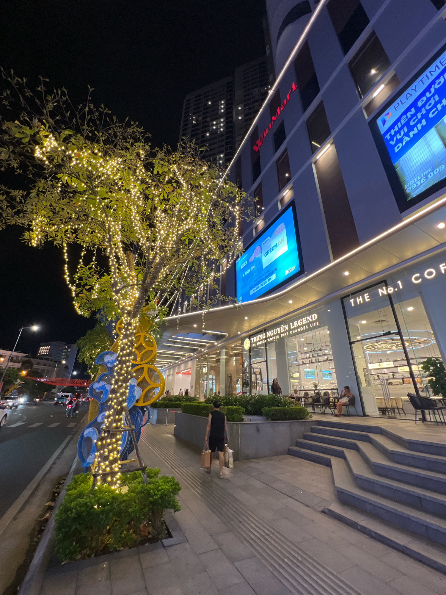 This is a nighttime photograph of a street in Nha Trang, Vietnam, specifically in front of a building with the "WinMart" logo. A tree adorned with numerous small, warm-toned lights is prominently featured on the left side of the image, casting a glow on the surrounding area. The building has several illuminated screens displaying advertisements, one of which shows "PLAY TIME THIÊN ĐƯỜNG VUI CHƠI DÀNH CHO". Below the screens, there is a storefront with the text "TRUNG NGUYEN LEGEND" and "THE ENERGY COFFEE THAT CHANGES LIFE". Seated at tables outside this establishment are a few individuals. On the sidewalk, a person wearing a black outfit is walking away from the camera, carrying shopping bags. The street is paved with grey tiles, and a set of wide, stepped stairs is visible on the right. Traffic lights and vehicles can be seen on the street in the background, indicating urban activity. The overall scene suggests a vibrant commercial area at night.