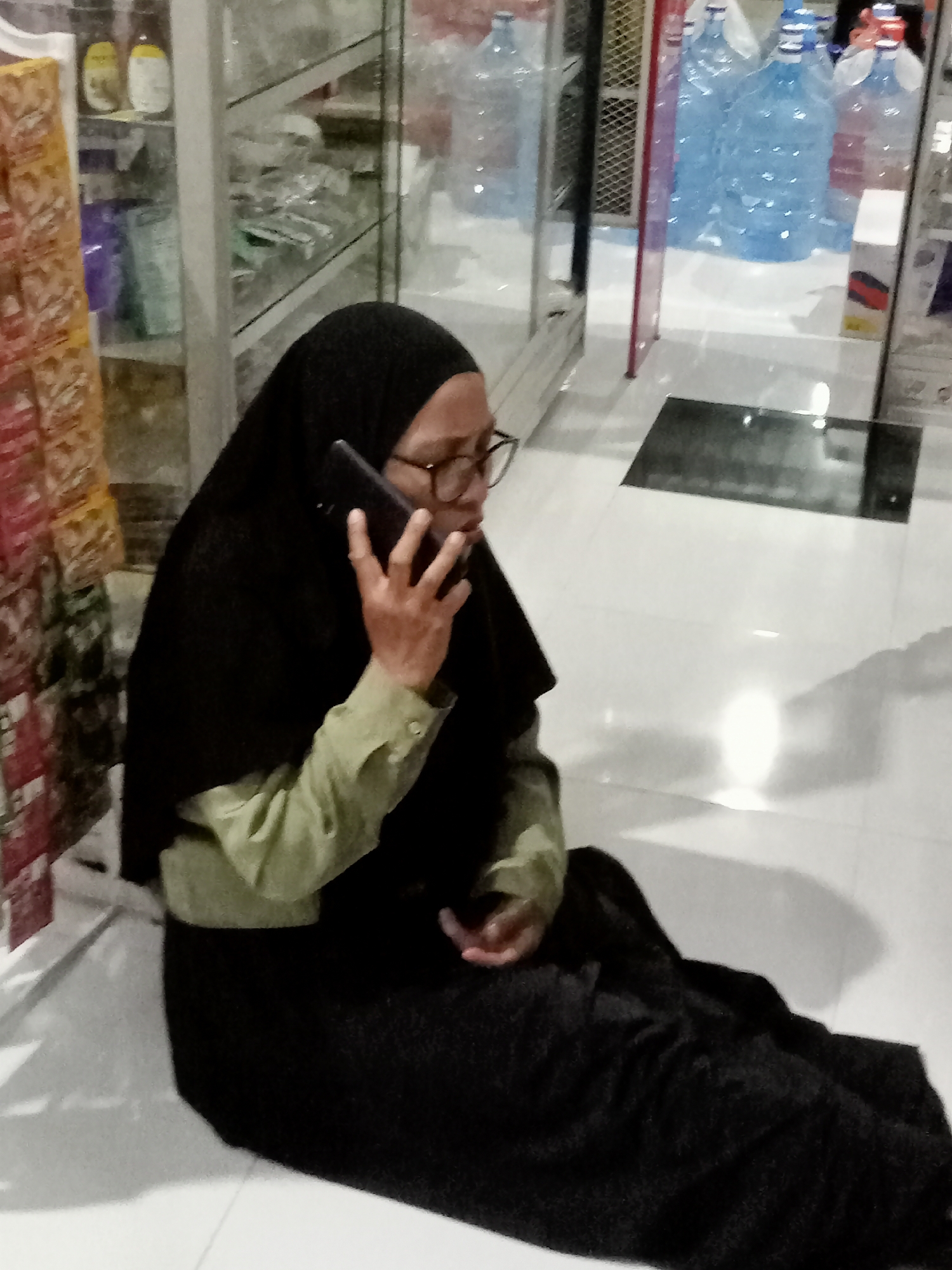 In Simo, Indonesia, a person wearing a black hijab and glasses is sitting on the floor, holding a black smartphone to their ear and speaking. They are wearing a light green long-sleeved shirt underneath their hijab. The background shows shelves stocked with various items, including bottles of water and packaged goods. The flooring appears to be white tiled. The lighting suggests an indoor environment, possibly a store or a small shop.