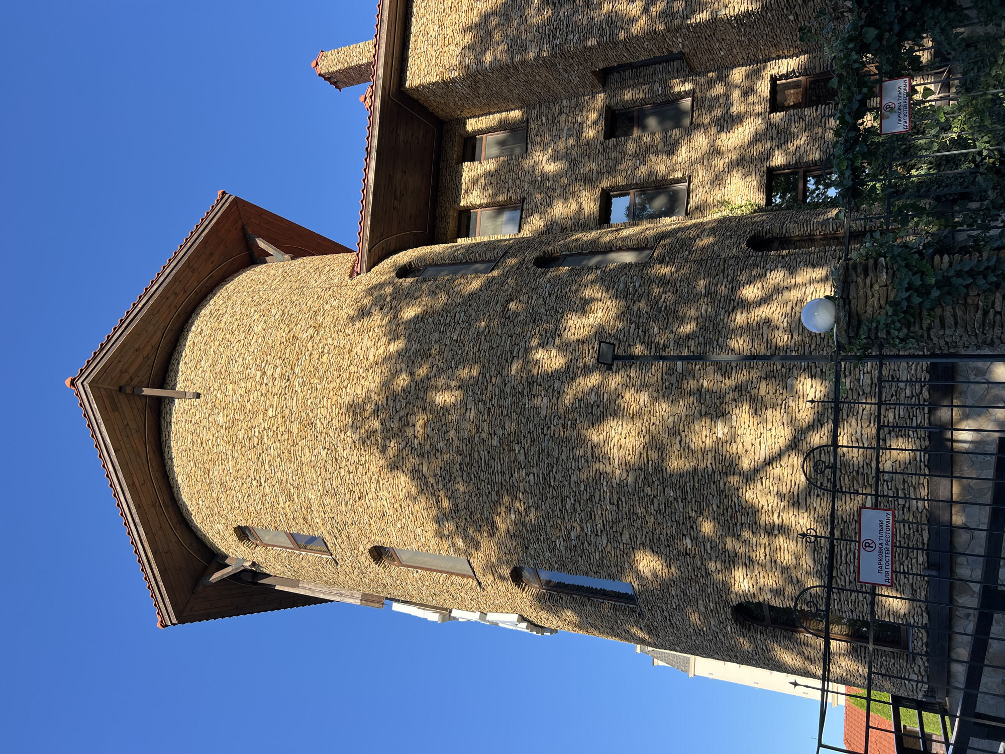 The image shows a stone tower-like building with a conical wooden roof, set against a clear blue sky. The building's exterior is covered in small, irregular tan-colored stones. Several windows with dark frames are visible on the tower, some arched and others rectangular. A section of the building to the right of the tower also features similar stonework and multiple windows. Overhanging wooden eaves are present, with some visible wooden beams supporting them. A red-tiled roof is visible, as is a stone chimney on the right side.
In the foreground, a black metal fence runs across the bottom of the image. Attached to the fence is a rectangular sign with a red border. The sign contains a red circle with a diagonal line through it, and the Cyrillic text "ПАРКОВКА ТІЛЬКИ ДЛЯ ГОСТЕЙ РЕСТОРАНУ" (Parking only for restaurant guests). Green ivy climbs on the fence and on a stone pillar to the left of the sign. A white spherical light fixture is mounted on a stone pillar to the left of the fence. The building appears to be illuminated by sunlight, casting dappled shadows from unseen trees onto the stone facade.
The scene is situated outdoors, with the bright sky indicating daylight. The architecture suggests a possibly historic or themed establishment, such as a restaurant or hotel, given the signage. While the specific location in Fontanka, Ukraine, is not visually represented in the image itself through landmarks or signage, the description is based on the visual elements present.