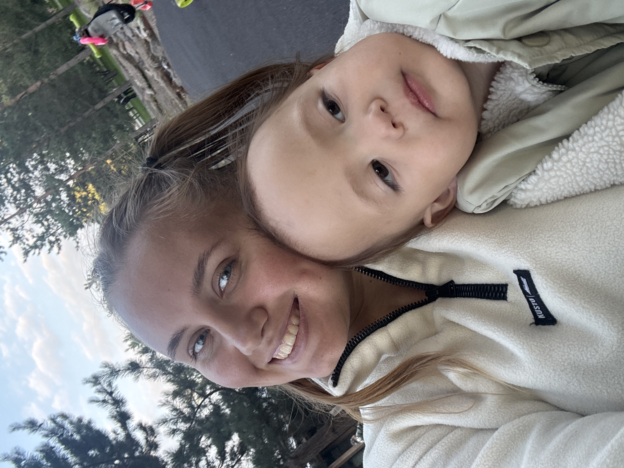 A close-up selfie shows a woman and a baby outdoors. The woman, positioned on the left, has blonde hair pulled into two pigtails and is smiling broadly, revealing her teeth. She is wearing a cream-colored fleece jacket with a black zipper. To her right, the baby is leaning against her, with a slightly pensive expression. The baby is wearing a light green jacket with a fuzzy white lining. The background suggests a park or outdoor recreational area, with trees and a paved surface visible. The lighting indicates it is daytime, possibly late afternoon given the soft light. The image appears to have been taken in the city of Krasnoyarsk, Russia, as indicated by the context provided.